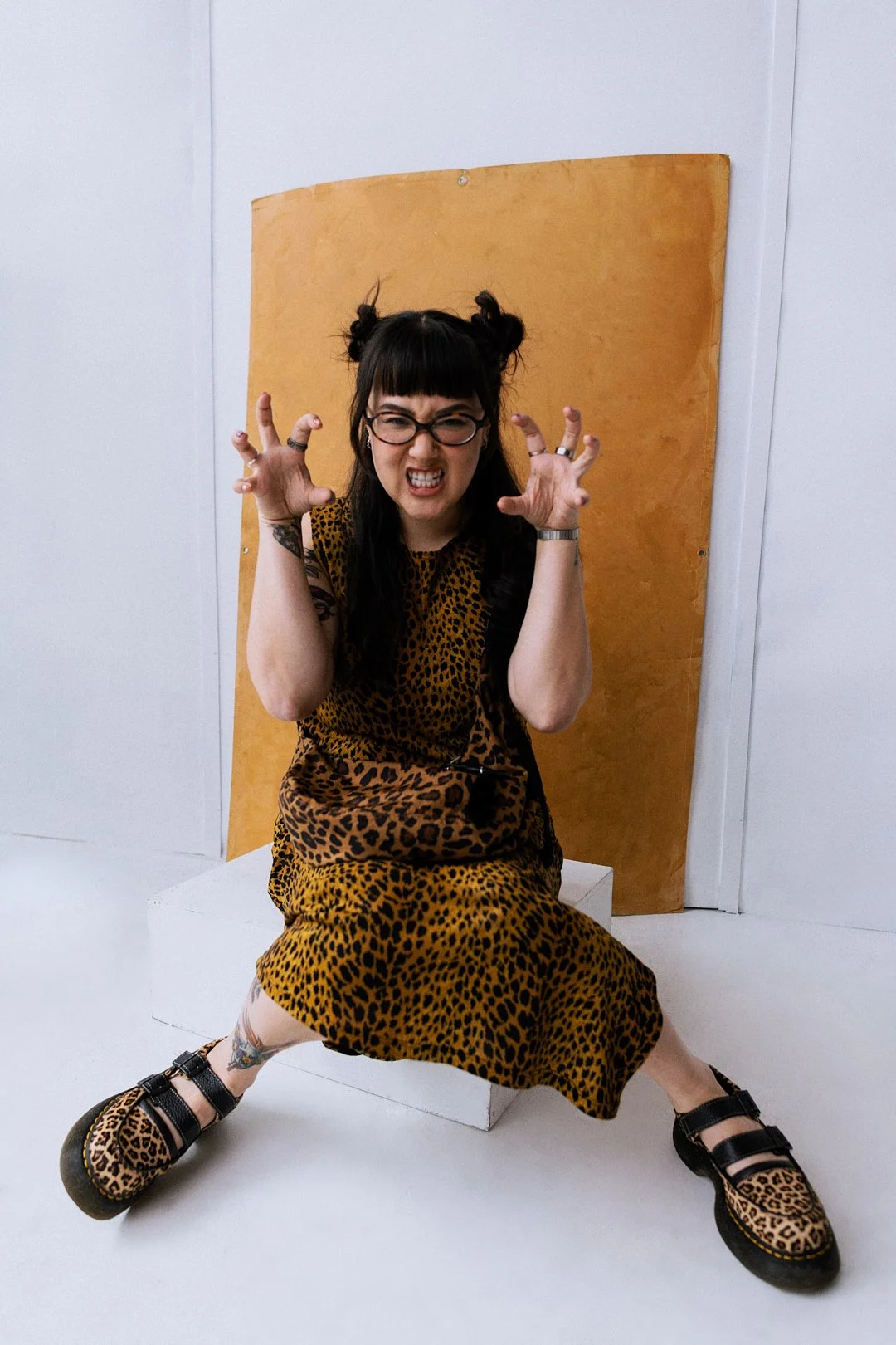 A woman with dark hair styled in buns, wearing glasses and a leopard print dress, sitting on a white block with a yellow background behind her, making a fierce expression and clawing her hands.