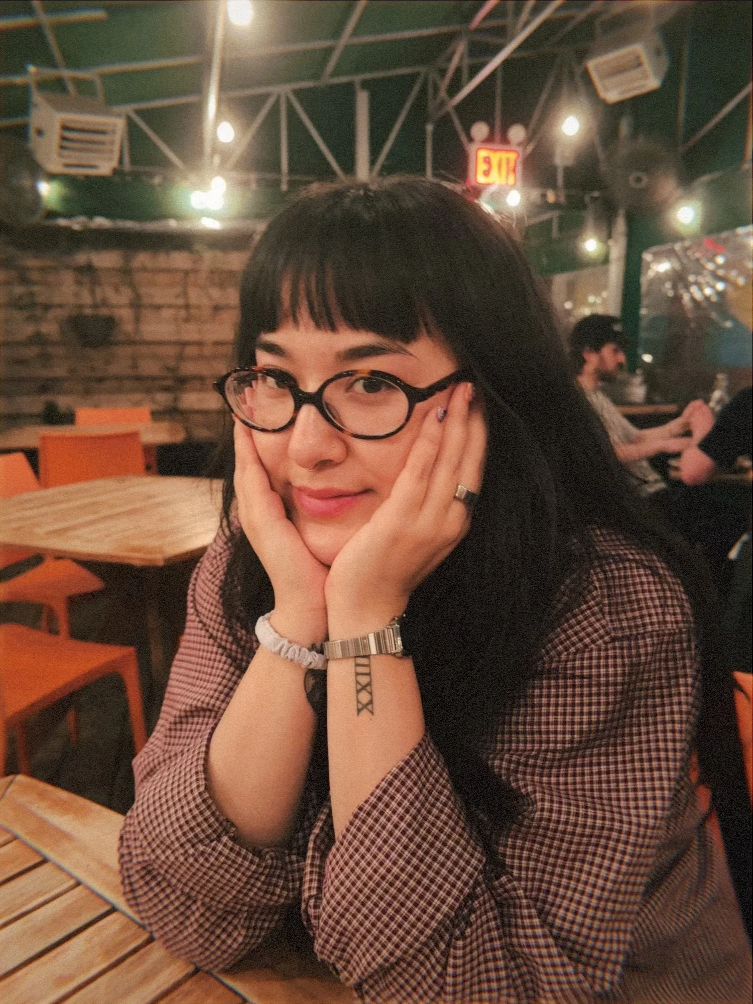 A young woman with dark hair, glasses, and a nose piercing, sitting at a restaurant table, resting her face in her hands, with a playful smile. She is wearing a checkered shirt, and her tattoos and accessories are visible on her arms. The background 