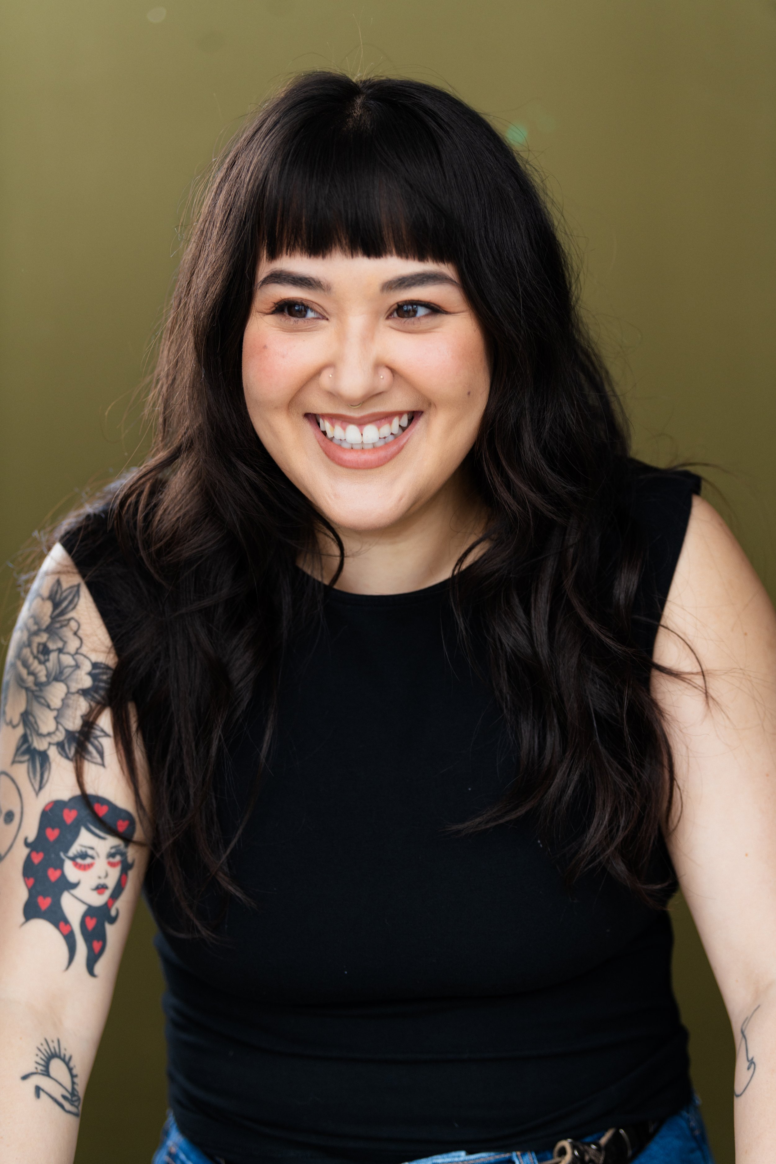 A woman with long dark hair, bangs, and tattoos on her arms, smiling, wearing a black sleeveless top, against a plain, light green background.