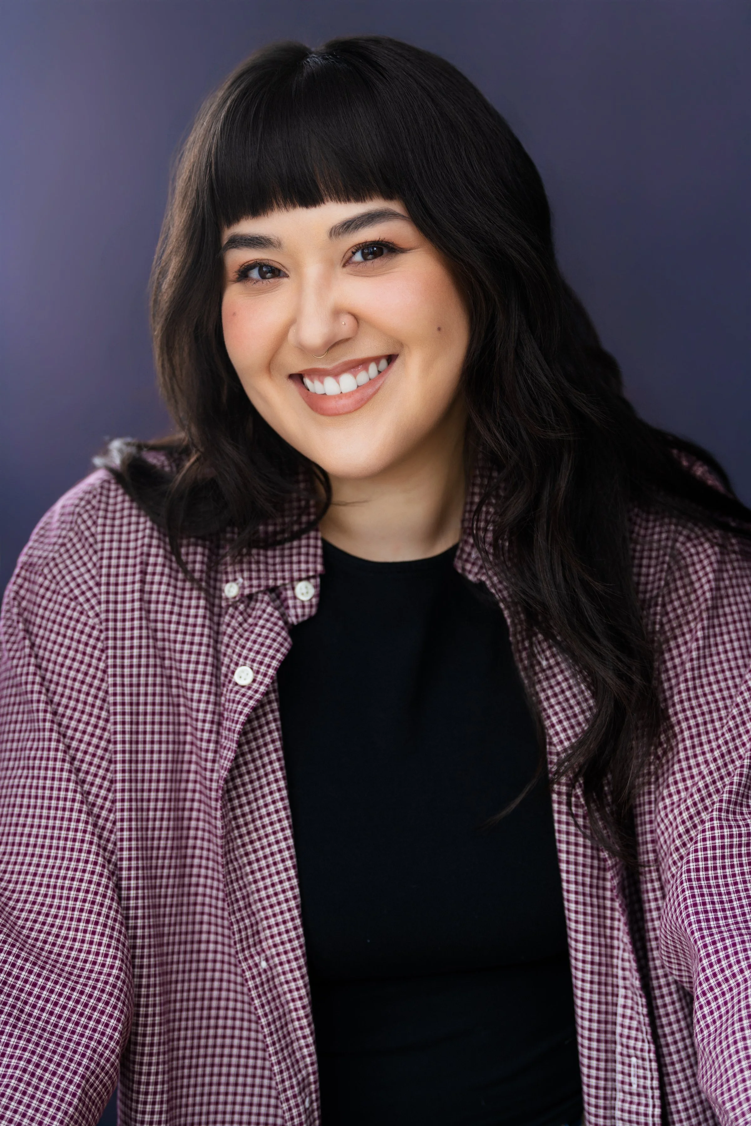 A young woman with black hair, bangs, and a nose piercing smiling at the camera, wearing a black top and a red and white checkered shirt, against a dark purple background.