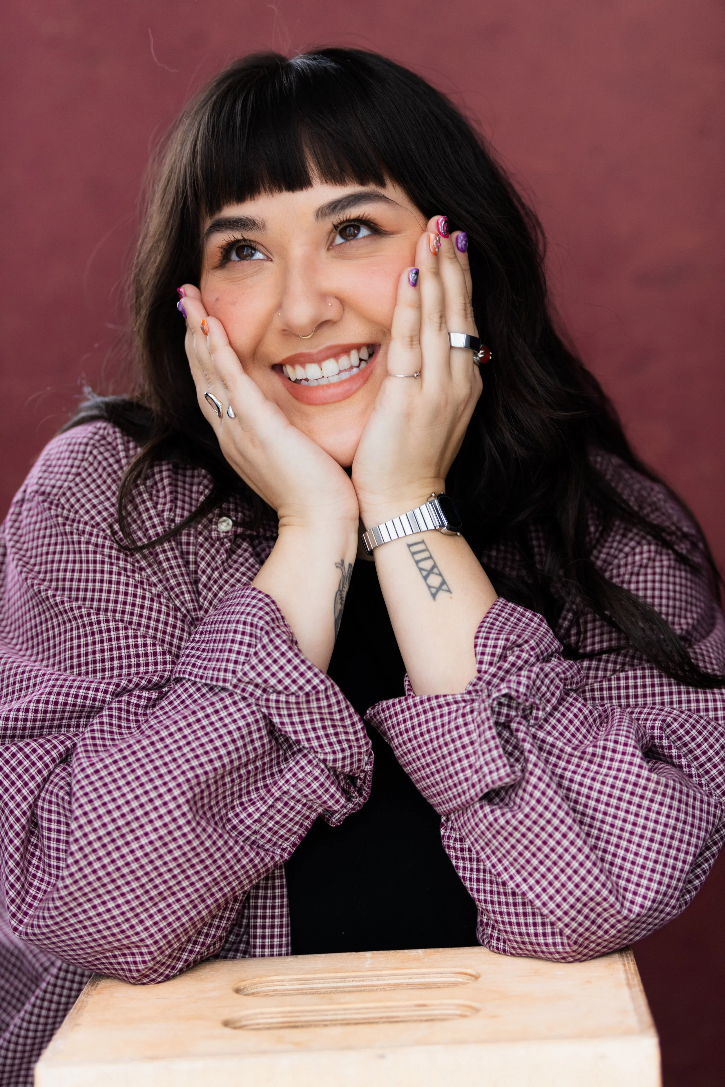 Young woman with dark hair and tattoos, smiling, resting her face in her hands, seated at a wooden table, wearing rings, bracelets, and a checkered shirt against a maroon background.