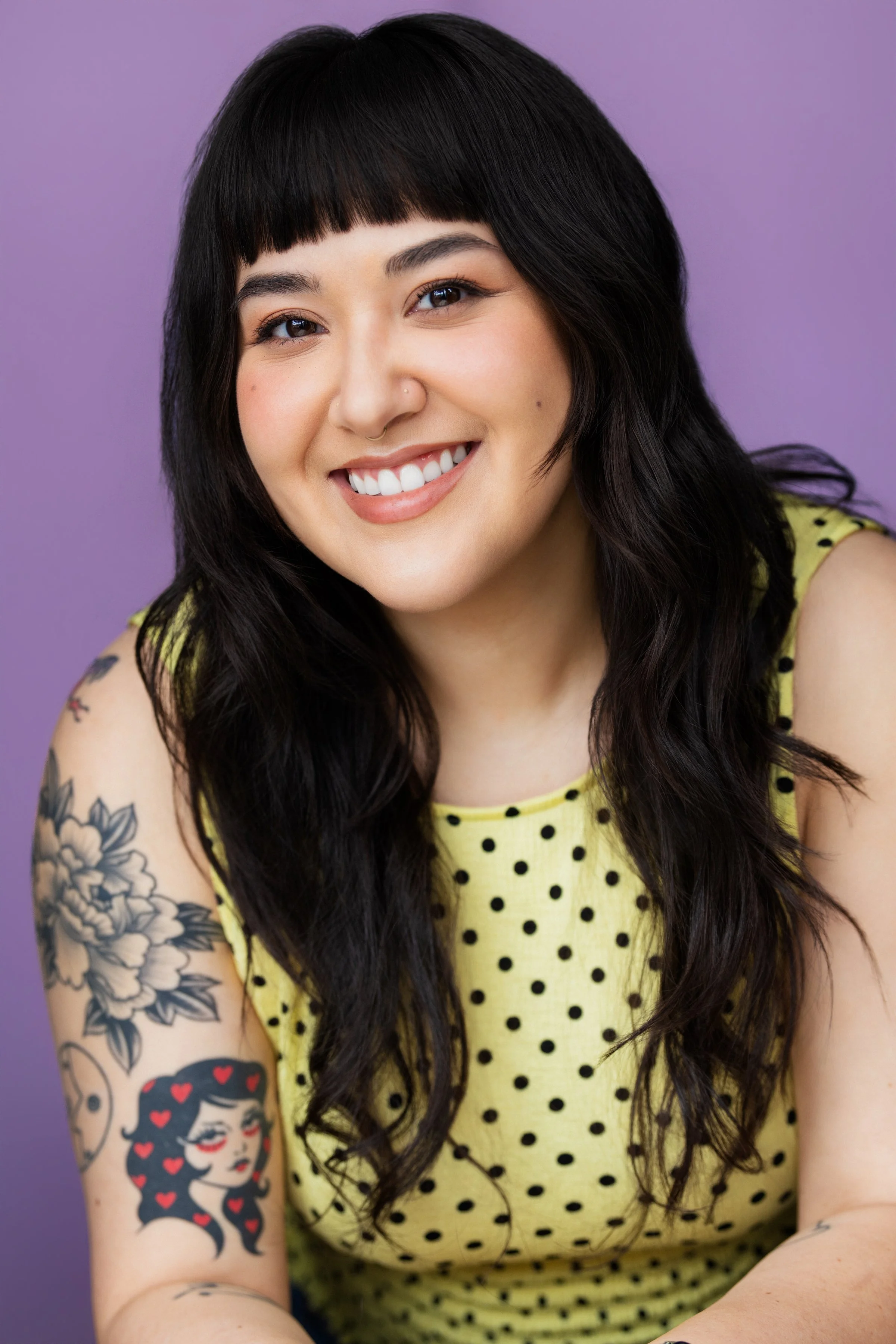 Close-up of a smiling woman with dark hair, a septum piercing, and colored tattoos on her arm, wearing a yellow dress with black polka dots, against a purple background.