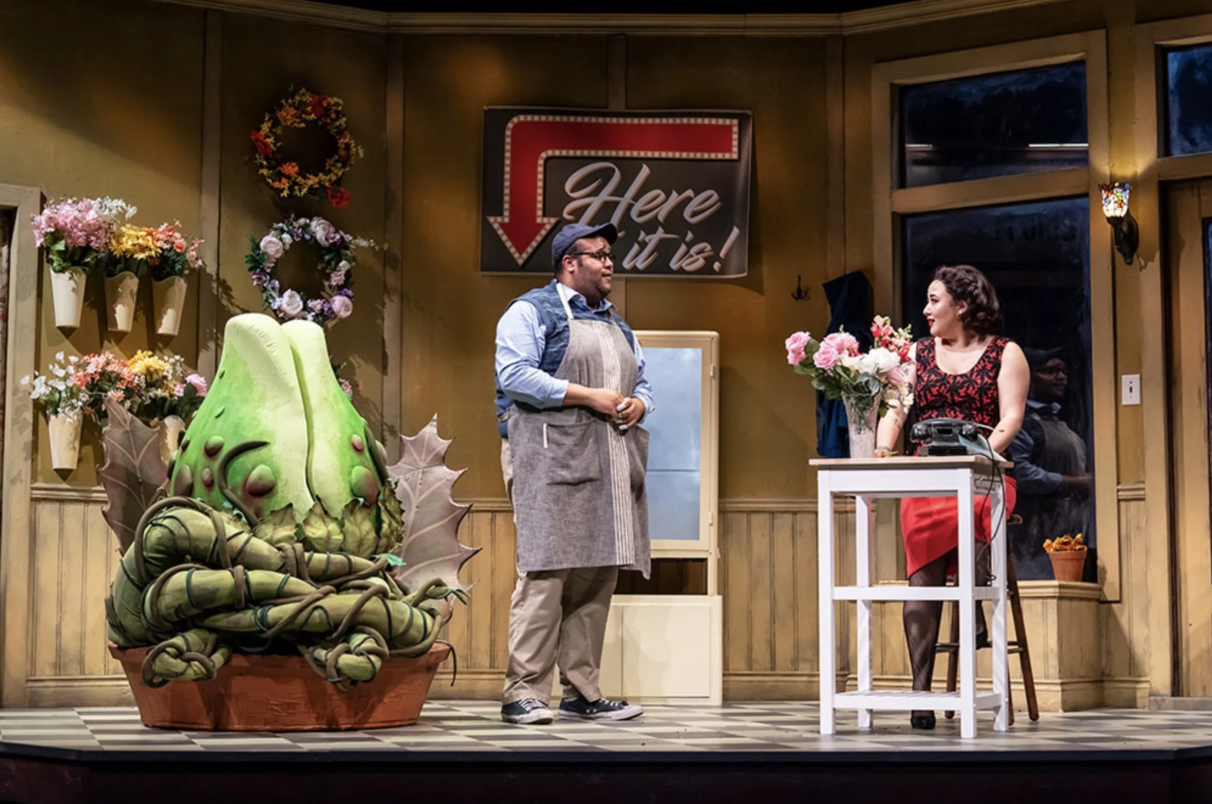 A scene from a theatrical play with two actors, one man and one woman, standing in a decorated room. The man is wearing glasses, a cap, a shirt, and an apron. The woman is dressed in a black and red dress, standing near a table with a vintage phone a