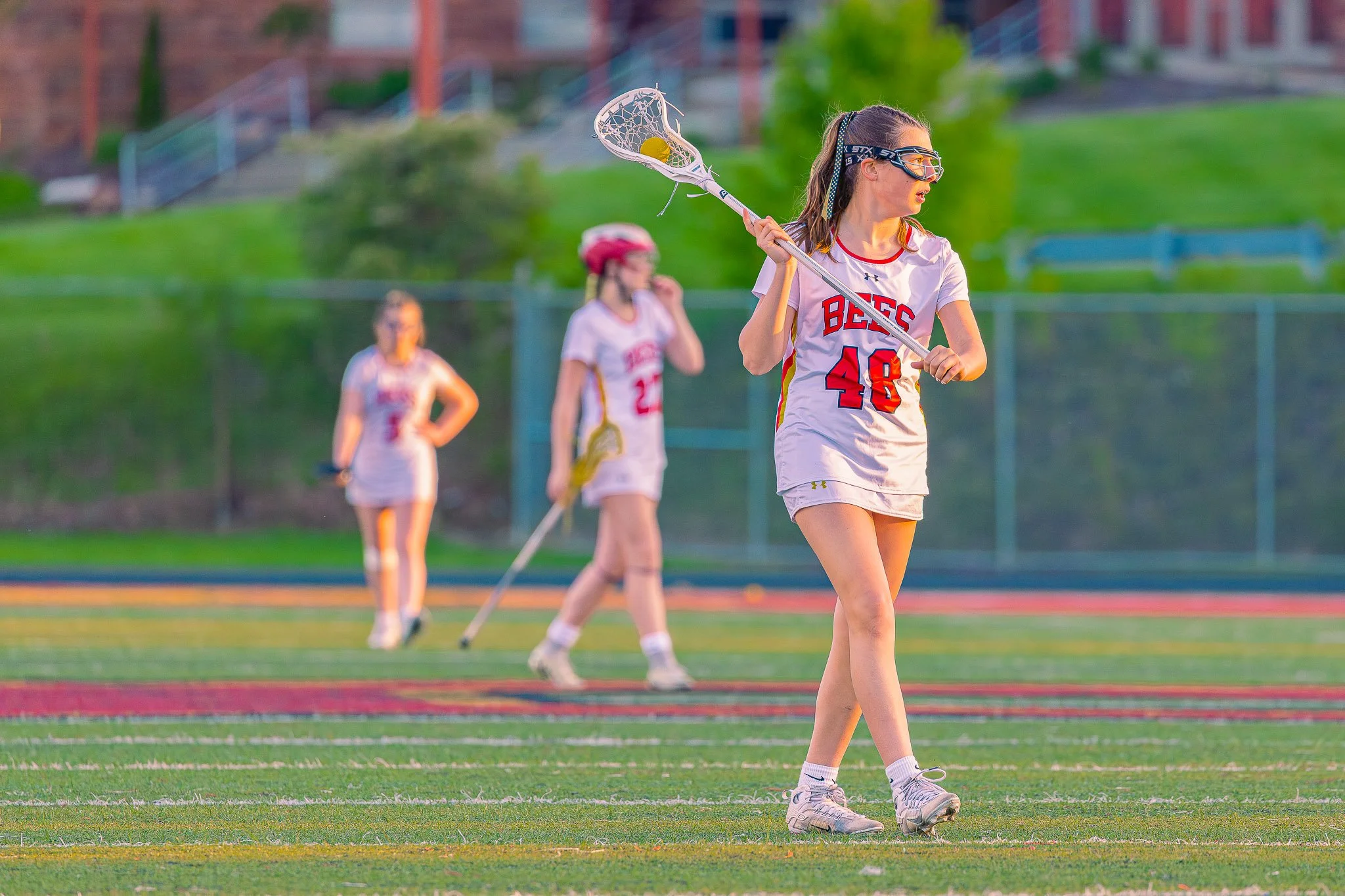 A girl playing lacrosse on a field, holding a lacrosse stick with a yellow ball, wearing sports goggles, a white uniform with red and yellow accents, and athletic shoes. Two other players can be seen in the background.