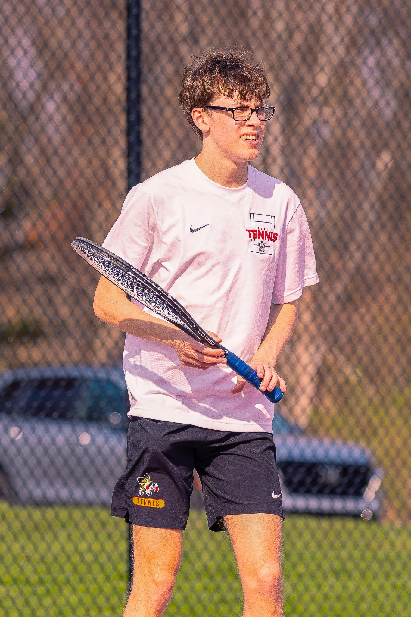 A young man with glasses and brown hair standing on a tennis court holding a tennis racquet.