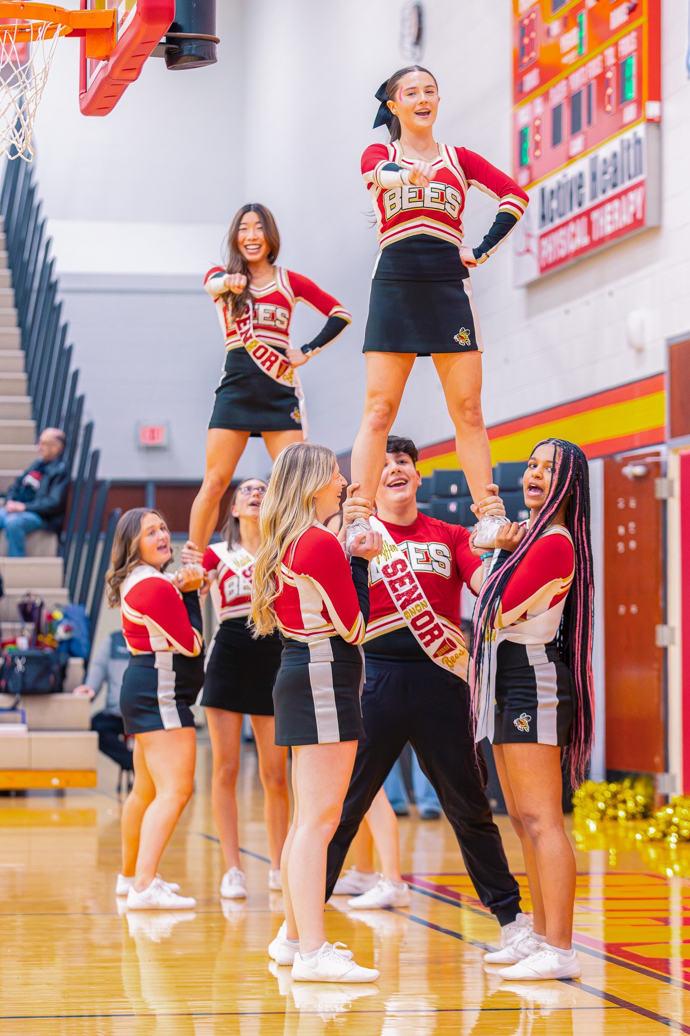 Cheerleaders performing a cheer routine indoors on a gymnasium floor, dressed in red, black, and white uniforms, with a basketball hoop and scoreboard in the background.