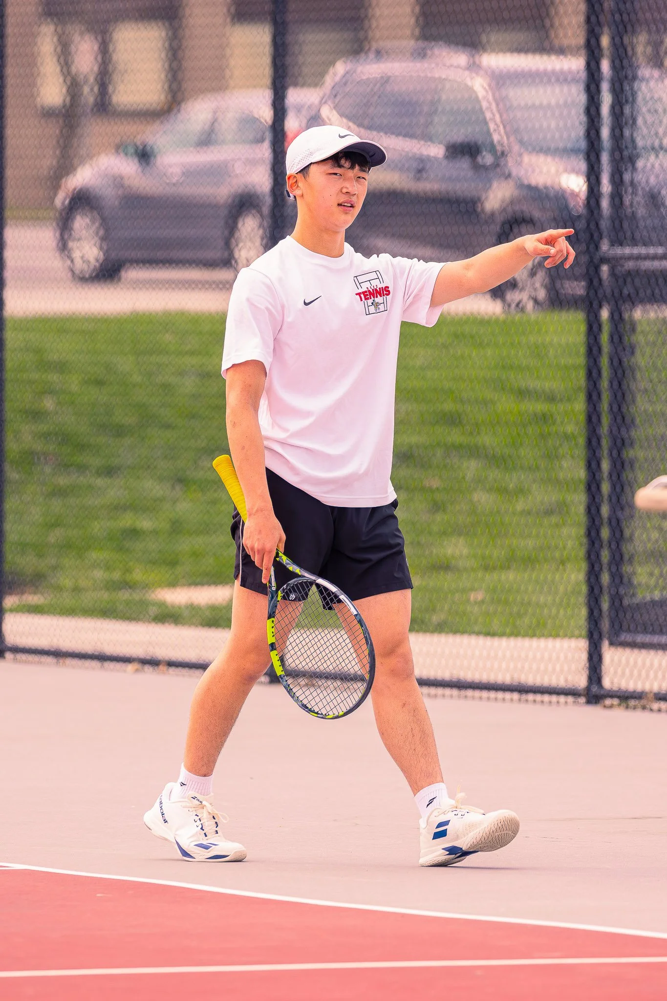 A young male tennis player on an outdoor court holding a tennis racket, wearing a white cap, white T-shirt with tennis logo, black shorts, and white tennis shoes, standing near the baseline, with a fence and parked cars in the background.