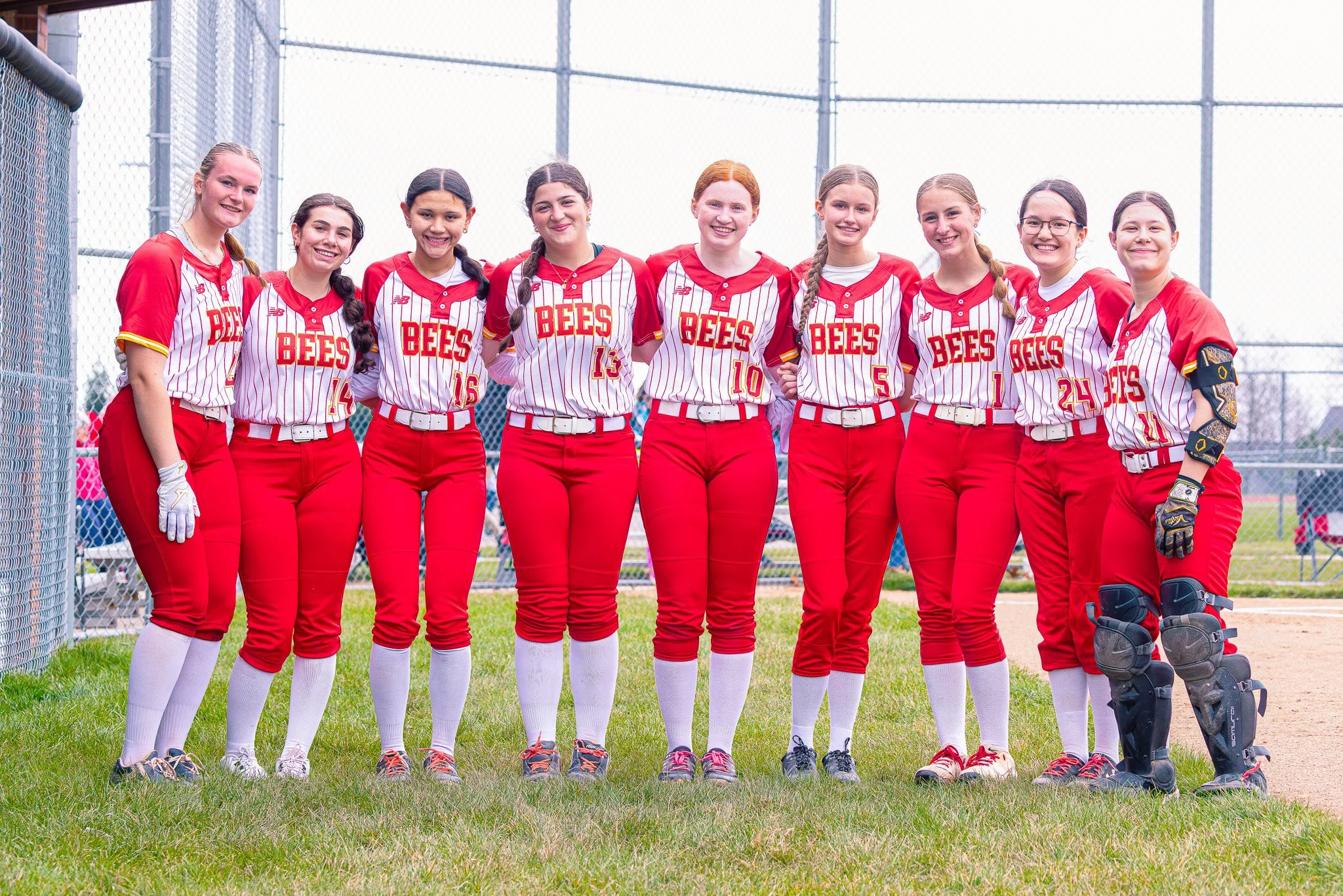 Young women baseball team standing together on the field in red and white uniforms, smiling at the camera.