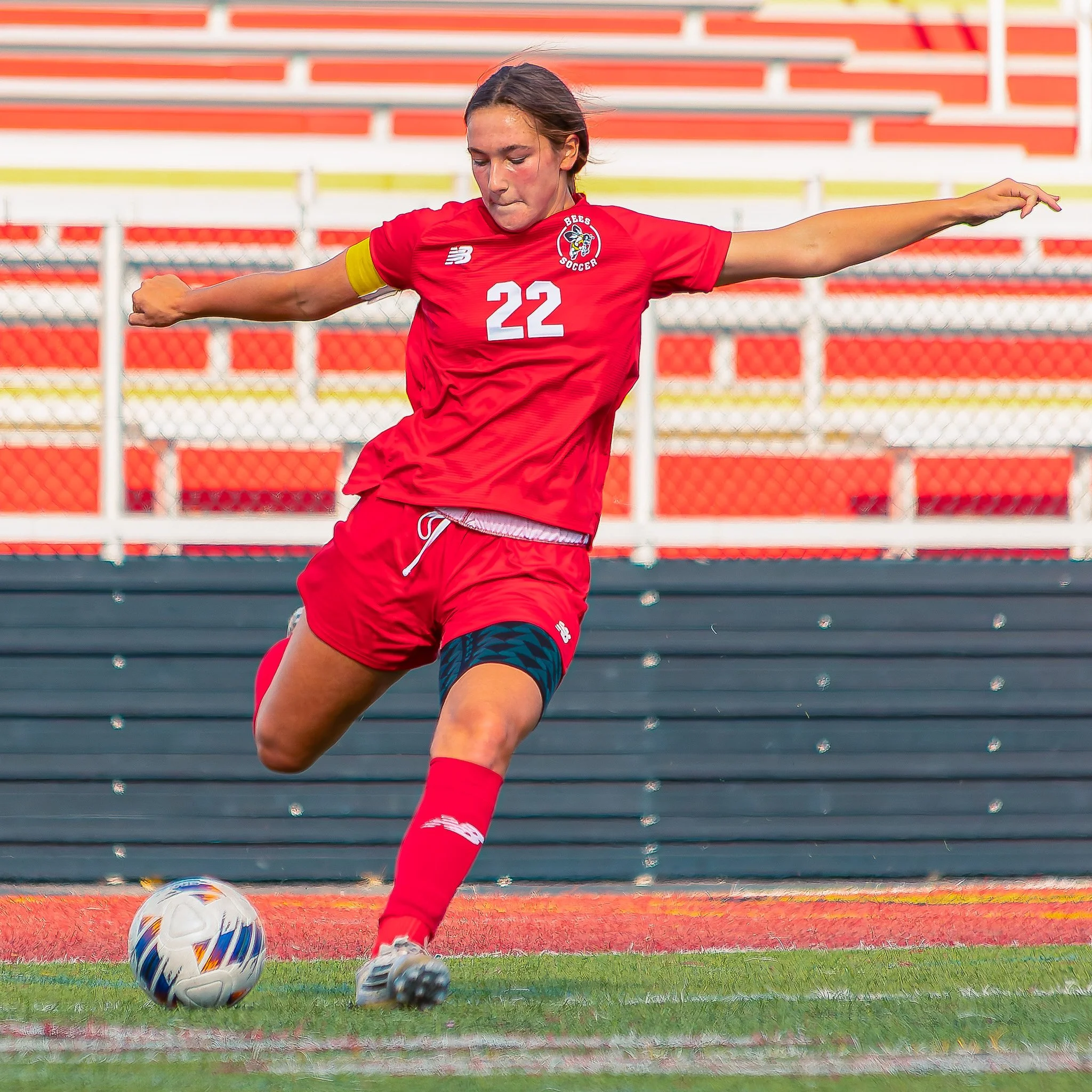 A female soccer player wearing a red jersey with the number 22 and a yellow captain's armband kicking a white soccer ball on a field with orange and green markings, with empty orange and yellow bleachers in the background.