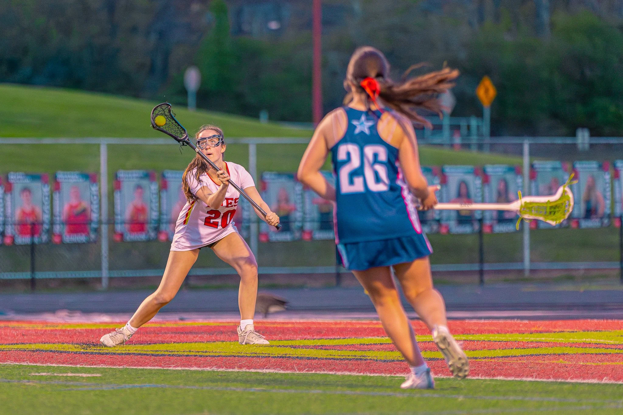 Two female lacrosse players are on the field, one in a pink jersey with number 20 holding a lacrosse stick, preparing to catch the ball, and the other in a blue jersey with number 26, wearing goggles and running with her lacrosse stick, during a game