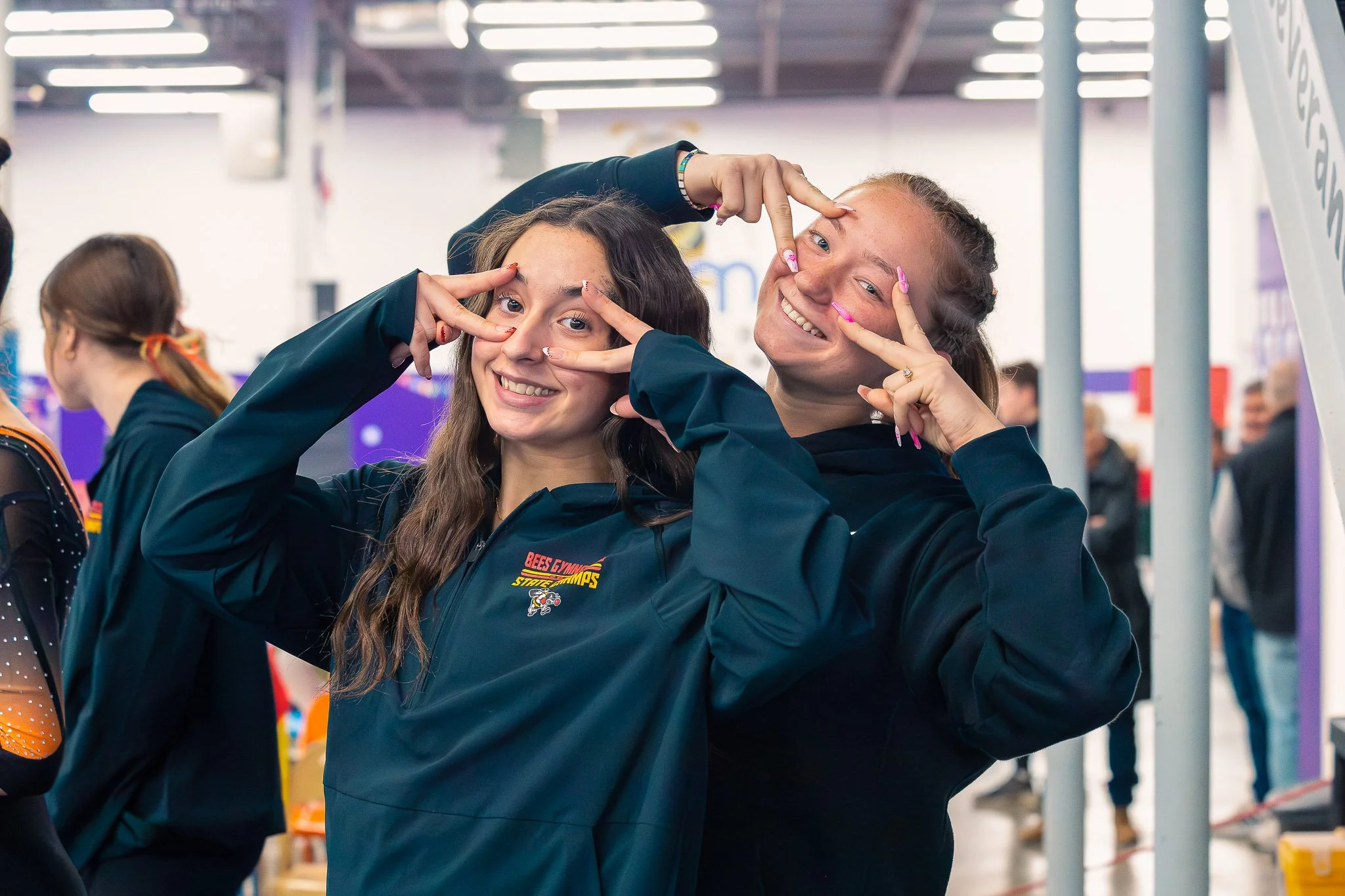 Two young women at an indoor event taking a selfie together, smiling and posing with peace signs, wearing matching dark jackets.