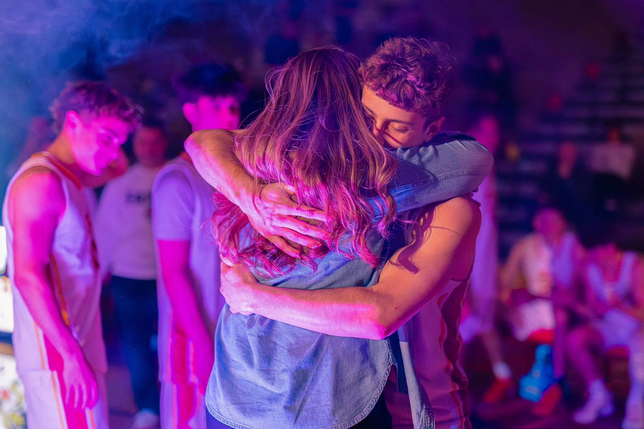 Two people hugging at a dance party or club with colorful lighting, surrounded by other people.