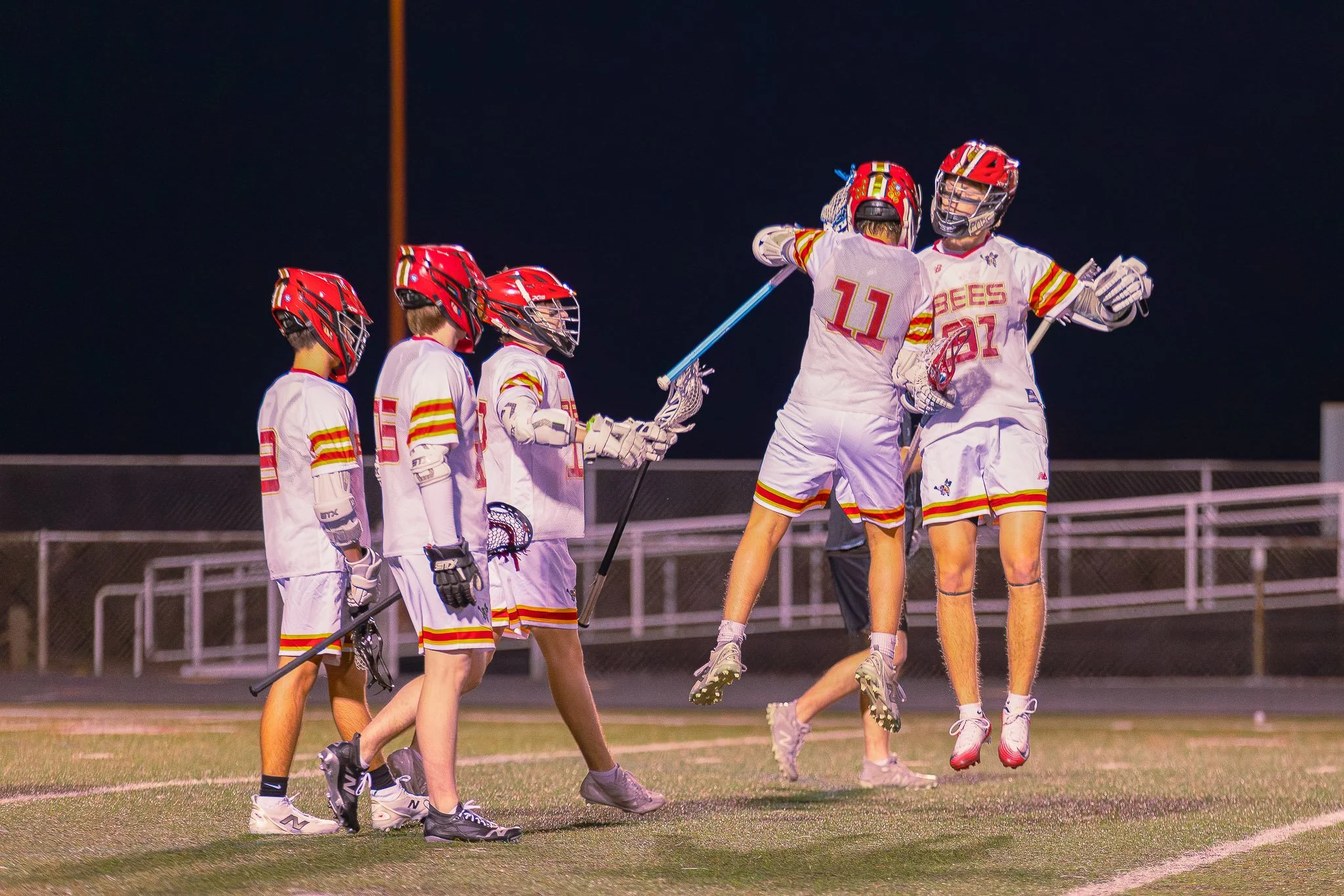 Lacrosse players in white uniforms and red helmets celebrating on the field at night.
