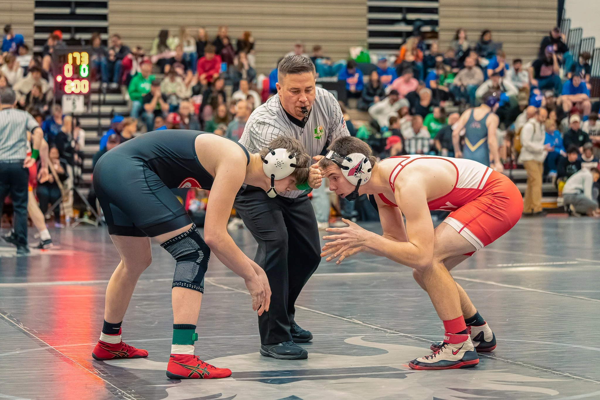 Two wrestlers, one in a black singlet and one in a red singlet, face off on a wrestling mat with a referee in a gray shirt overseeing the match. The referee is making a call while holding a whistle in his mouth. Spectators watch from bleachers in the