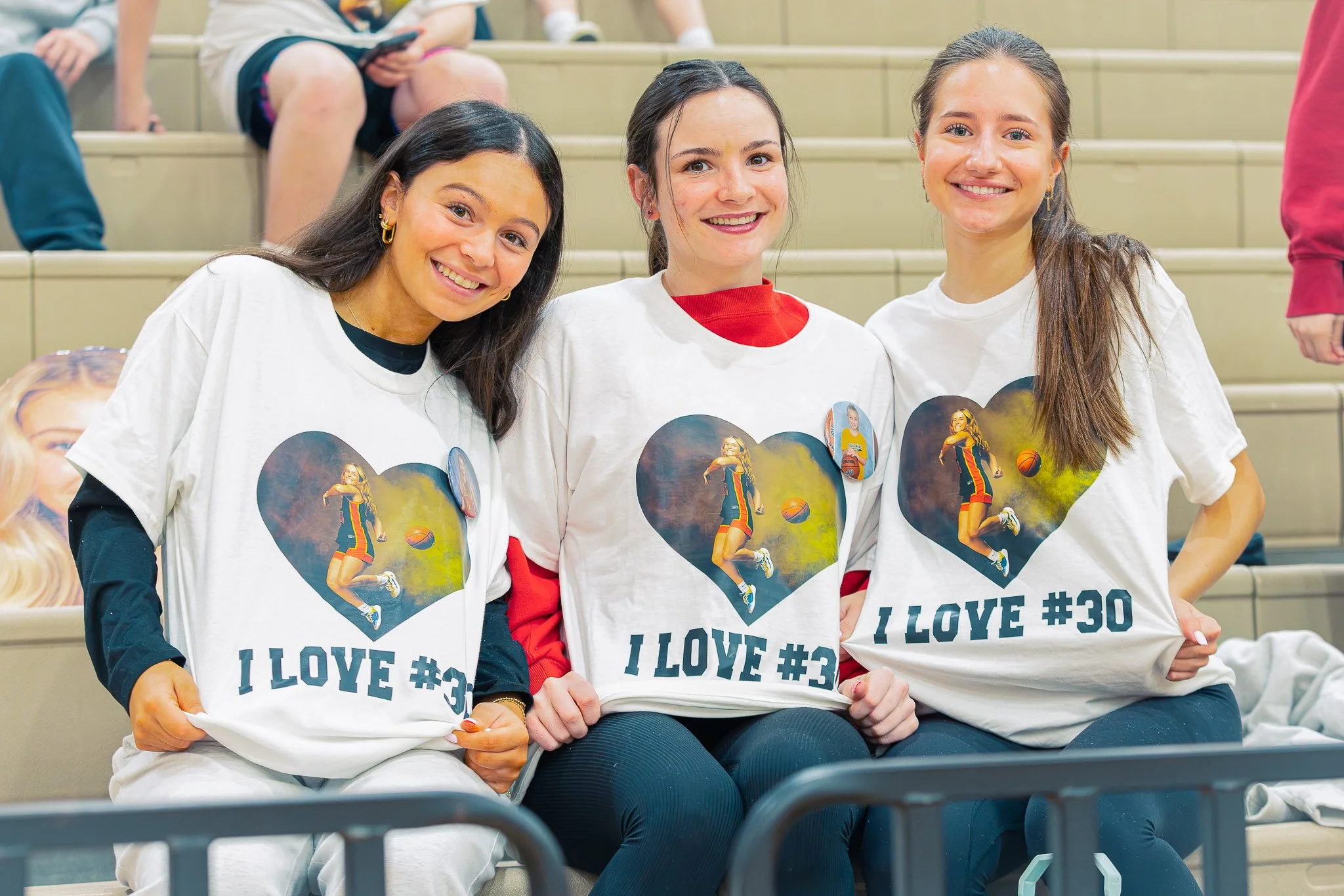 Three young women sitting in stadium seats, smiling, wearing white t-shirts with a basketball player on the front and the words "I LOVE #30".