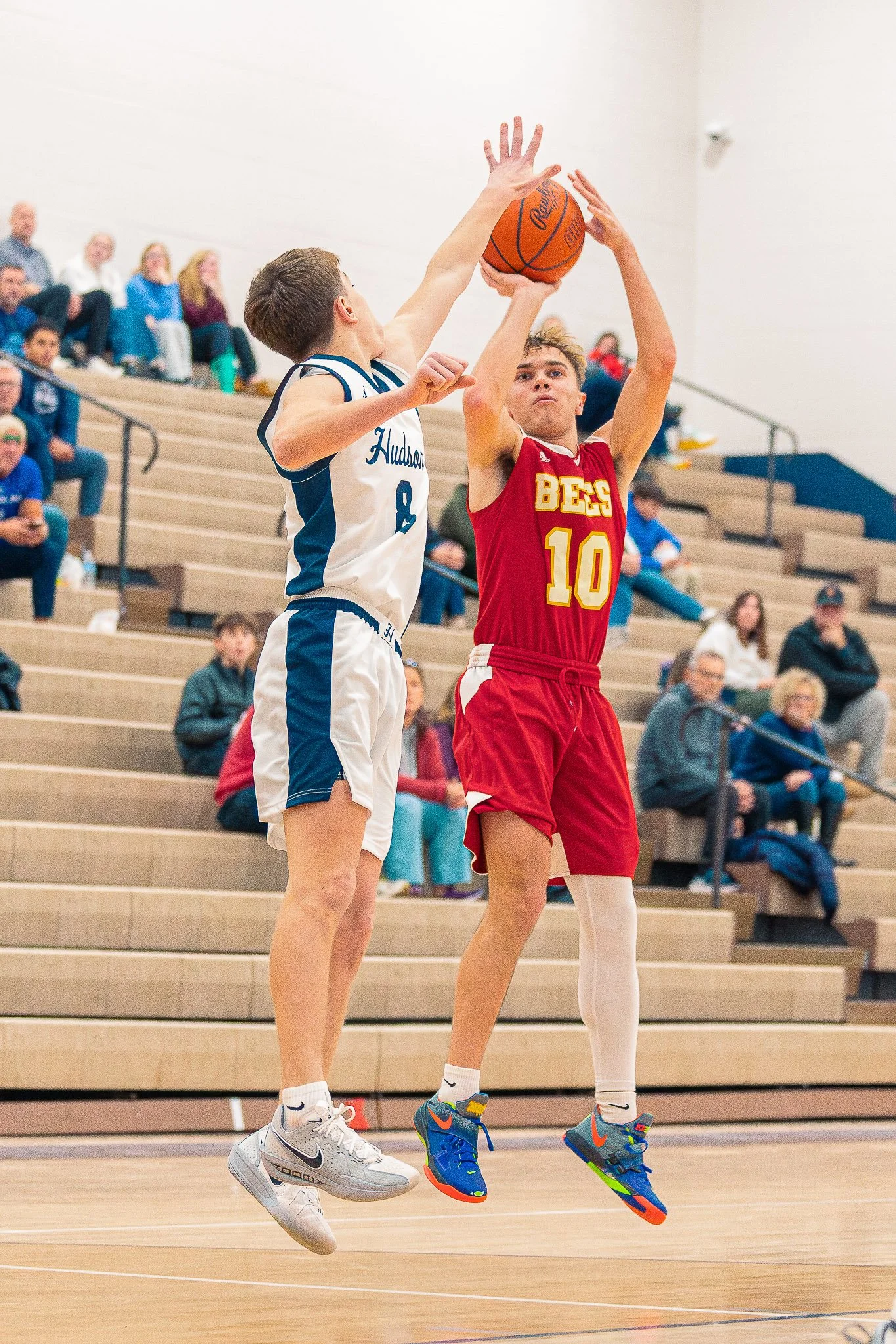 Two basketball players jumping for the ball during a game in an indoor gym, with spectators seated on wooden bleachers watching.