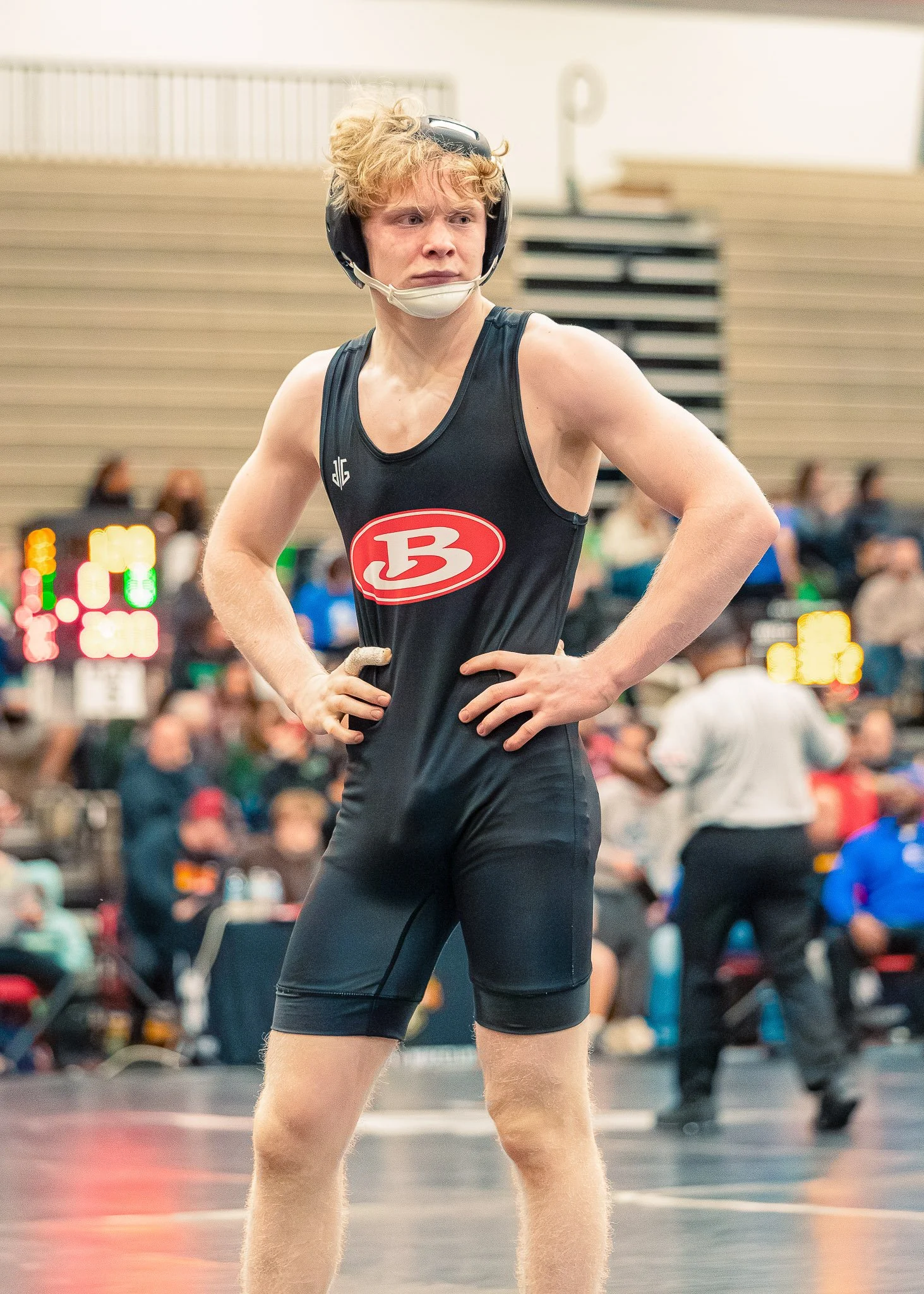 Young male athlete with curly blond hair, wearing a black sports singlet with a red logo, black compression shorts, a black helmet, and a face mask, standing in a gym or sports facility.