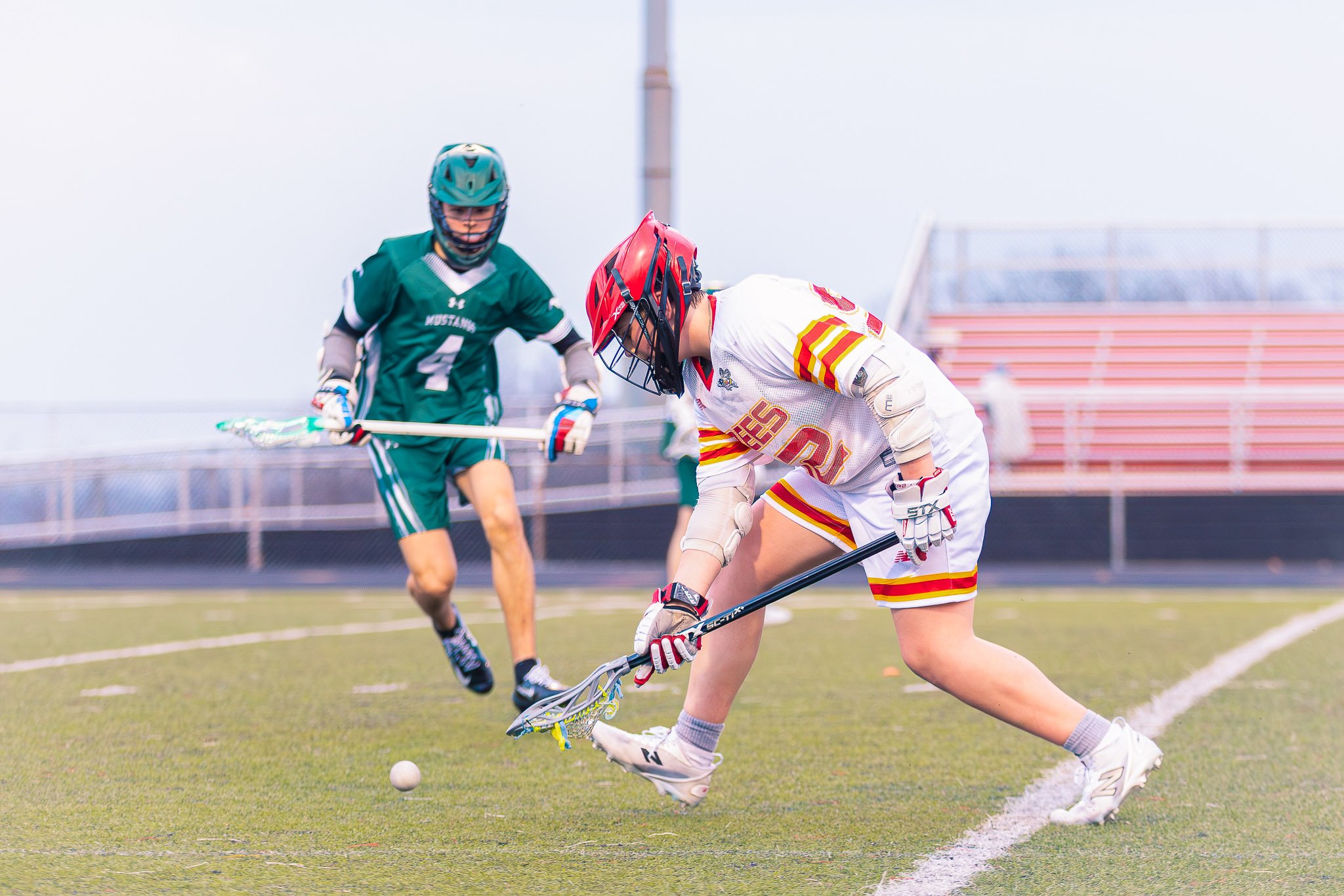 Two athletes in protective gear playing field hockey on a green turf field during daytime, with empty bleachers and a cloudy sky in the background.