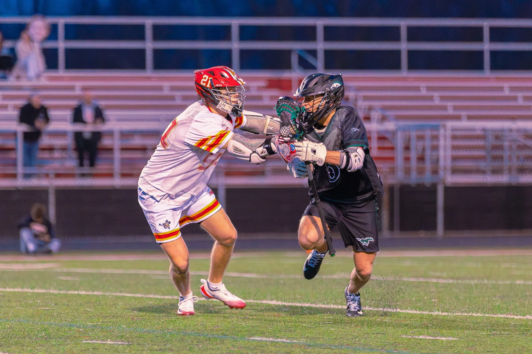 Two players are competing in a lacrosse game on a field. One player is wearing a white jersey with red and yellow accents and a red helmet, while the other player is dressed in black gear with a black helmet. The player in black is holding a lacrosse