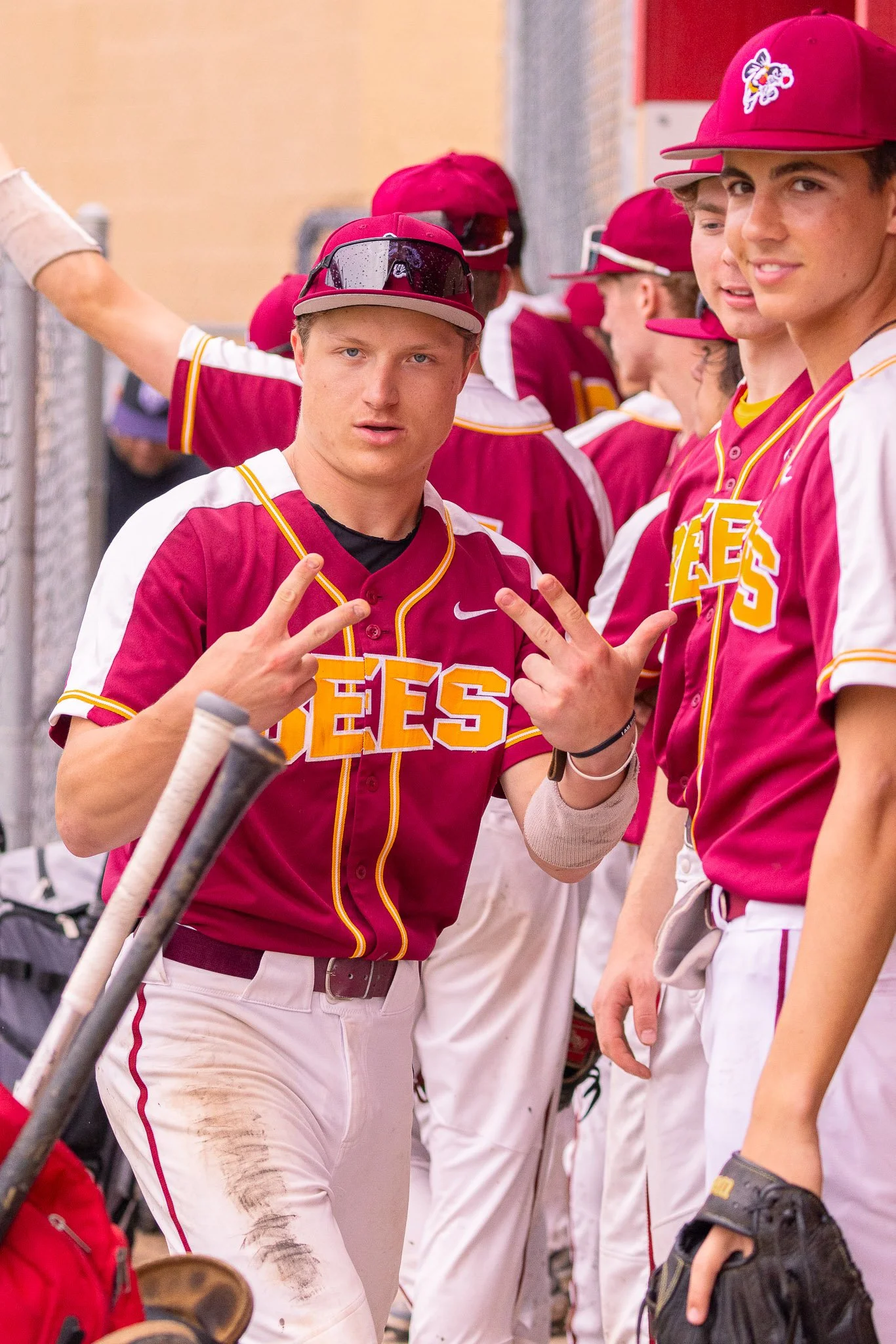A group of teenage baseball players wearing maroon and white uniforms with yellow accents, some wearing maroon caps with logo, gathered on a field. One player in foreground making a peace sign, holding baseball bats, some players smiling or talking, 