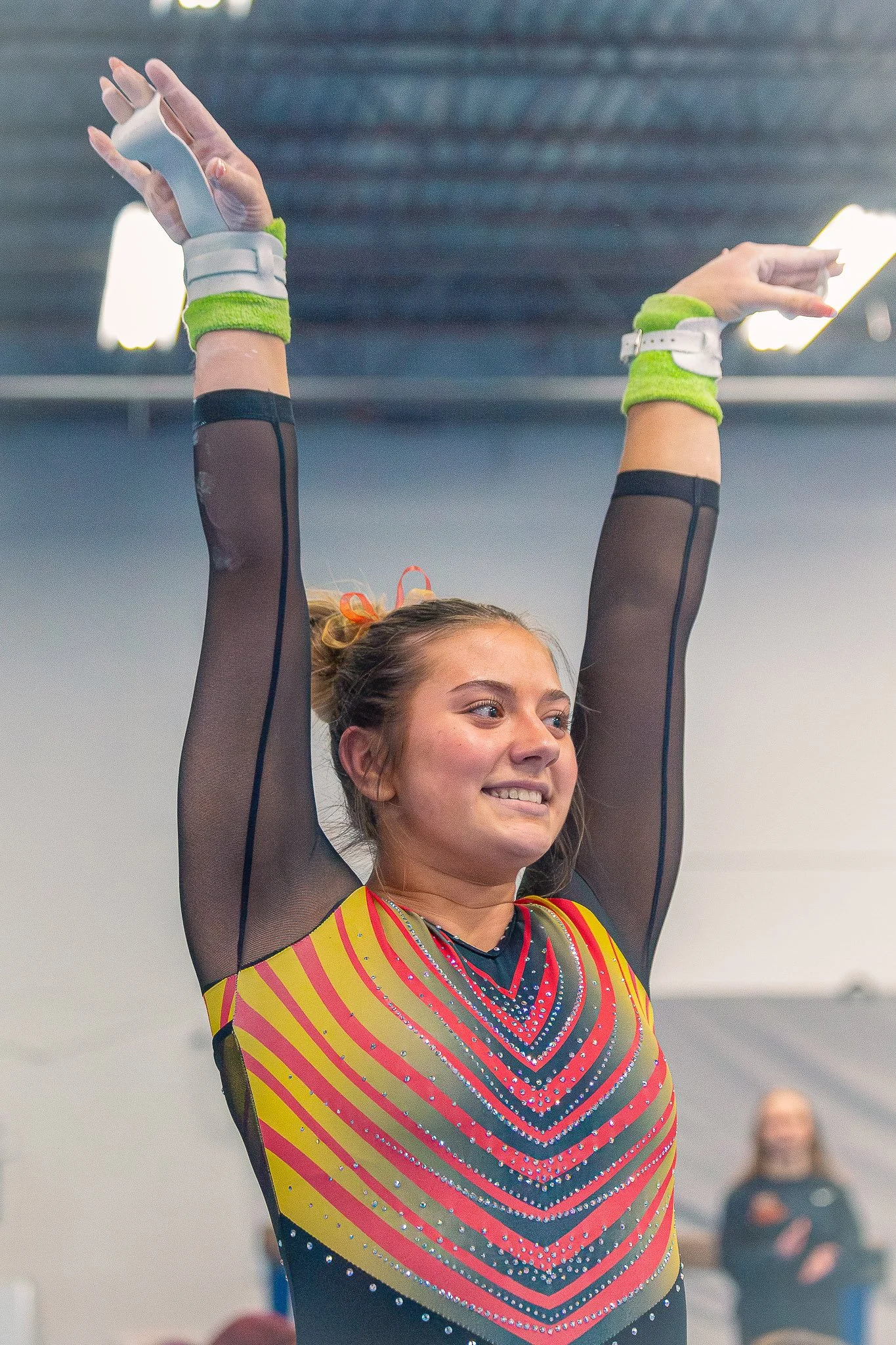 A young woman in a colorful, sparkly cheerleading uniform is smiling and raising her arms above her head. She appears to be in a gymnasium or sports arena with spectators in the background.