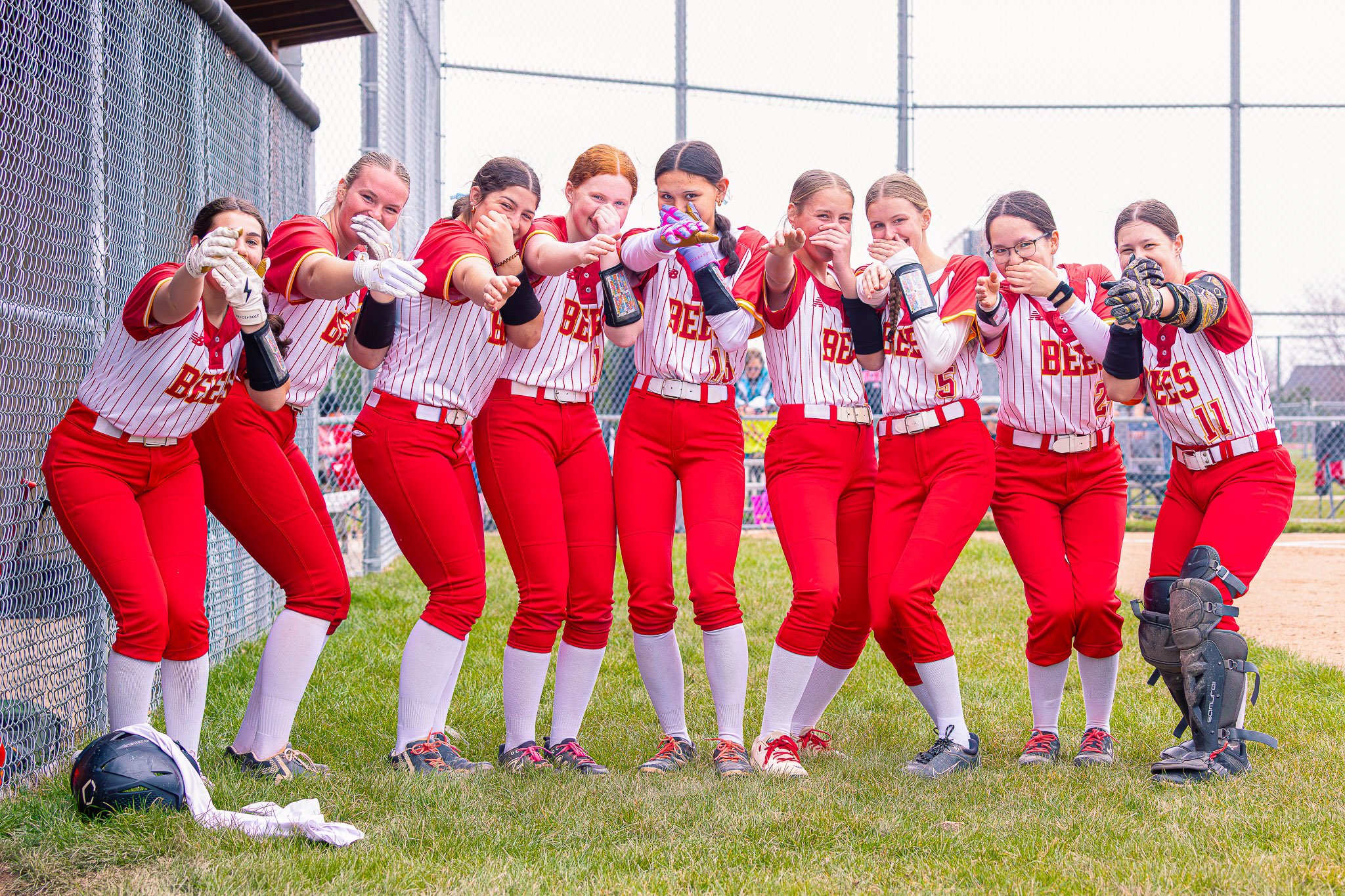Group of nine young female softball players wearing red and white uniforms, standing together near a chain-link fence on a softball field, some smiling and pointing at the camera.