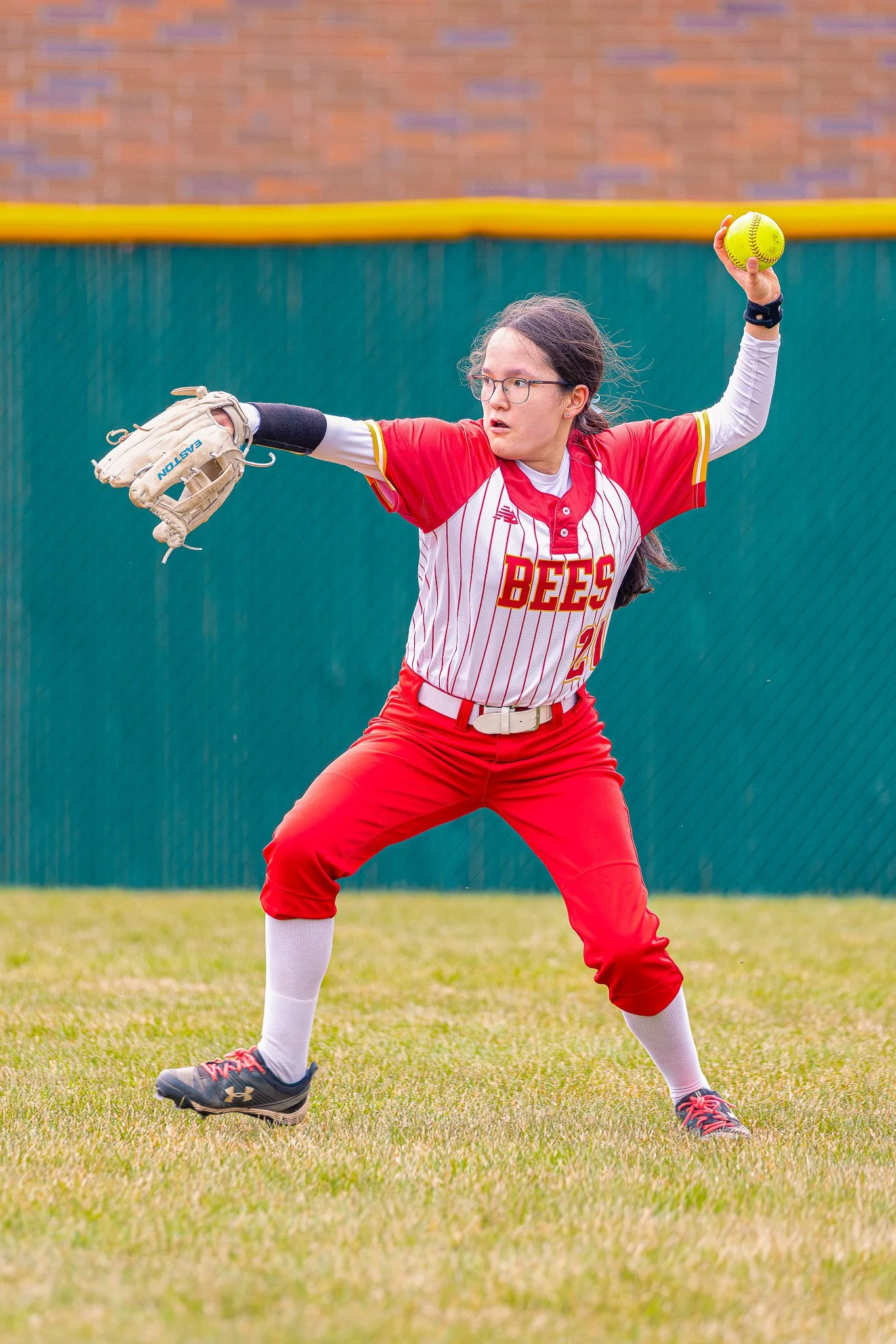 A female softball player in a red and white uniform with the word 'BEES' on the front, wearing glasses, and holding a yellow softball in her right hand. She appears to be in a ready position on the field.