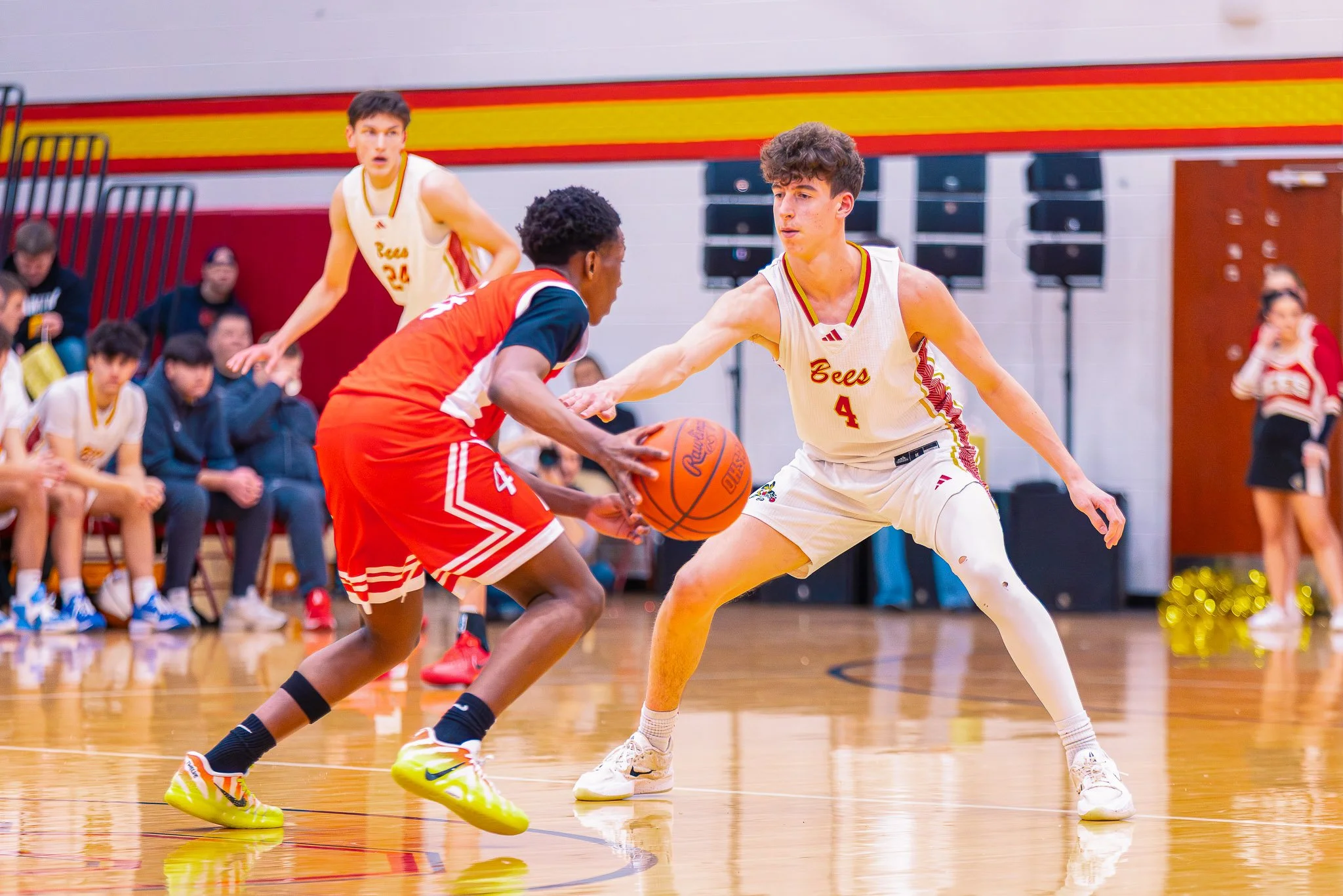 Two high school basketball players from opposing teams compete for the ball during a game in a gymnasium, with spectators seated on the sidelines.