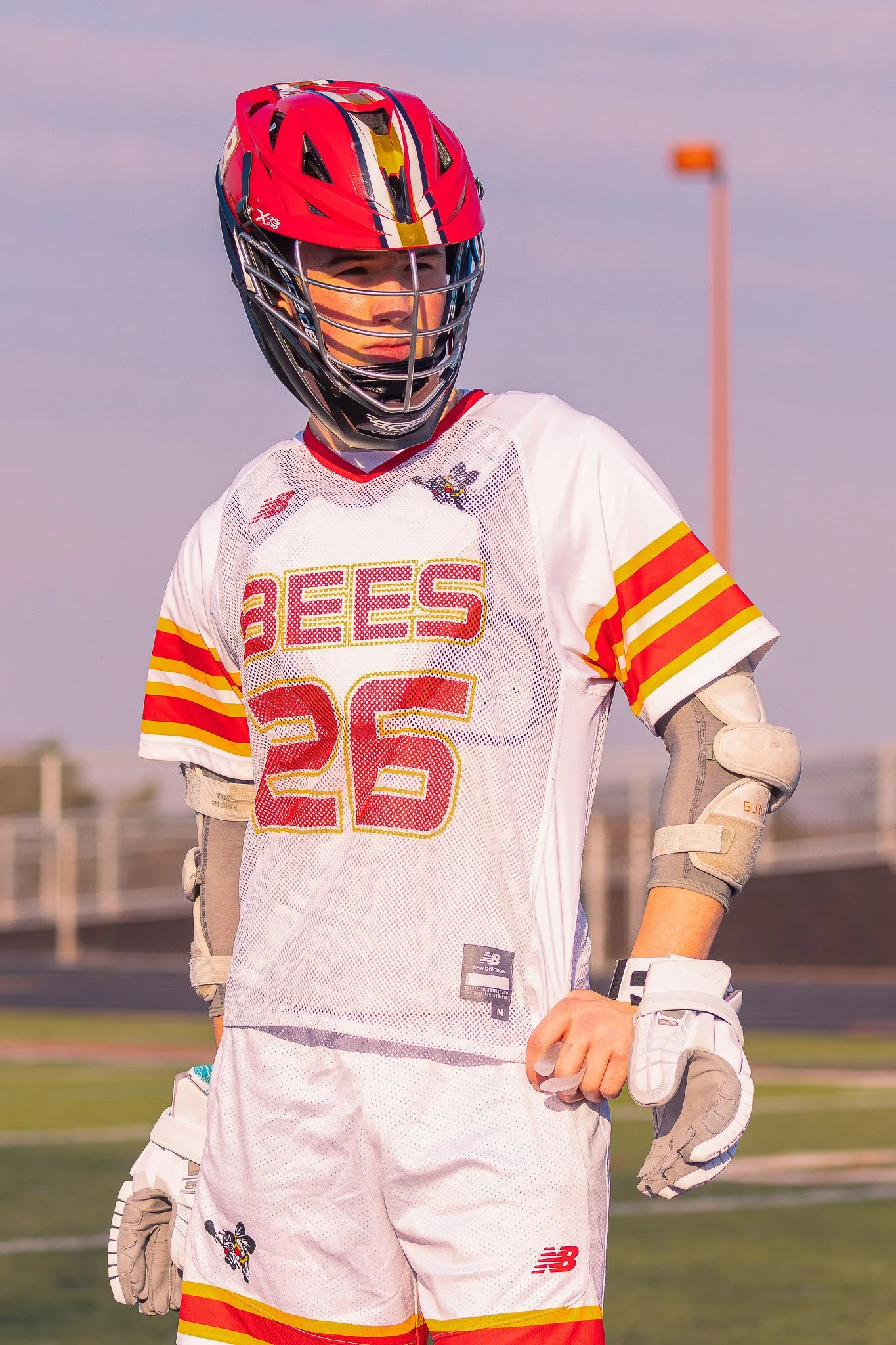 A young male lacrosse player in a red helmet with a face mask, wearing a white jersey with red and yellow stripes and the text 'BEES' with the number 26, standing on a sports field.