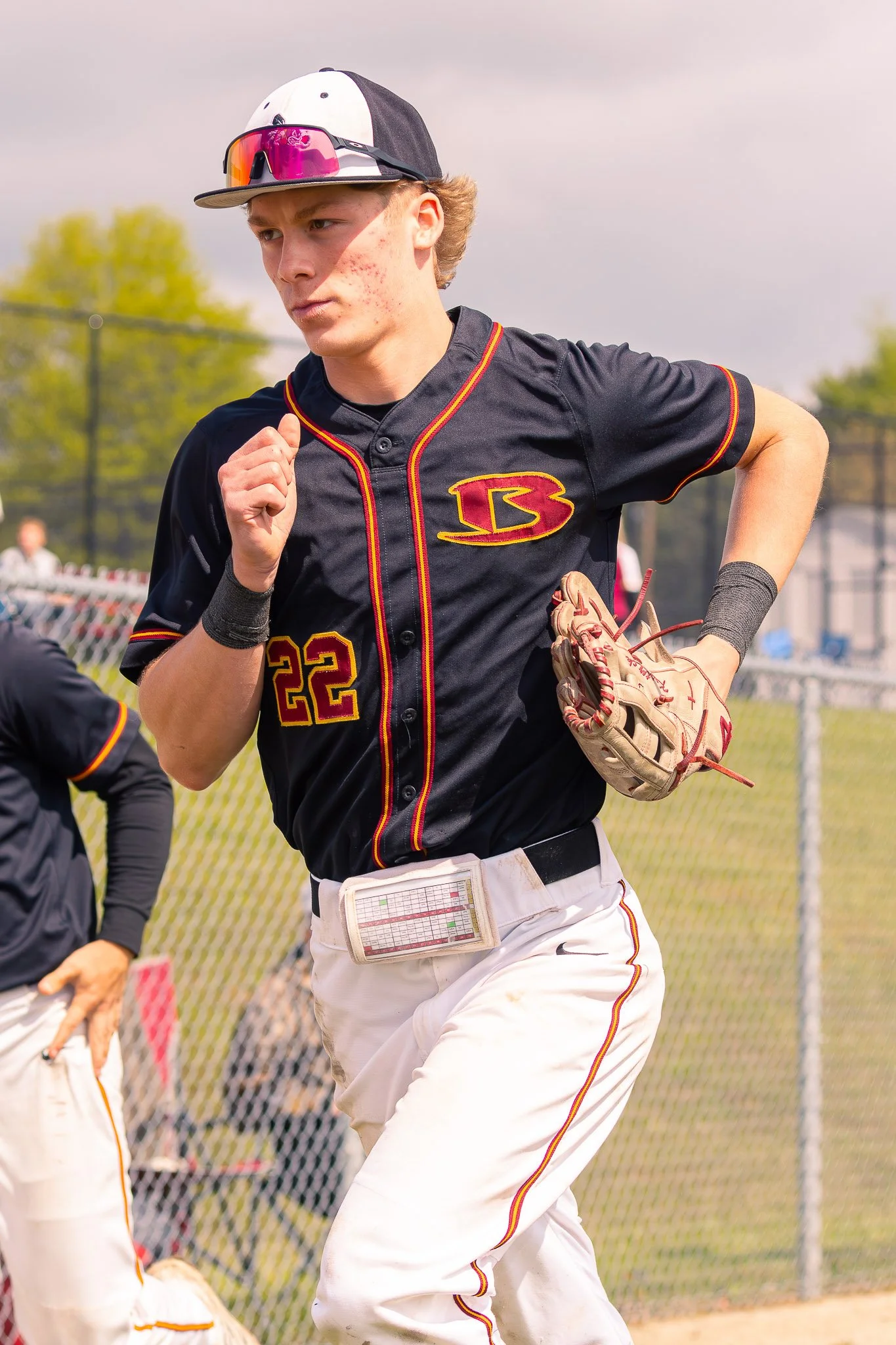 A young male baseball player with light skin, wearing a black baseball jersey with red and yellow accents, the number 22, and a logo on the chest. He has on a white and black cap with sunglasses on top and a glove on his left hand. He appears to be r