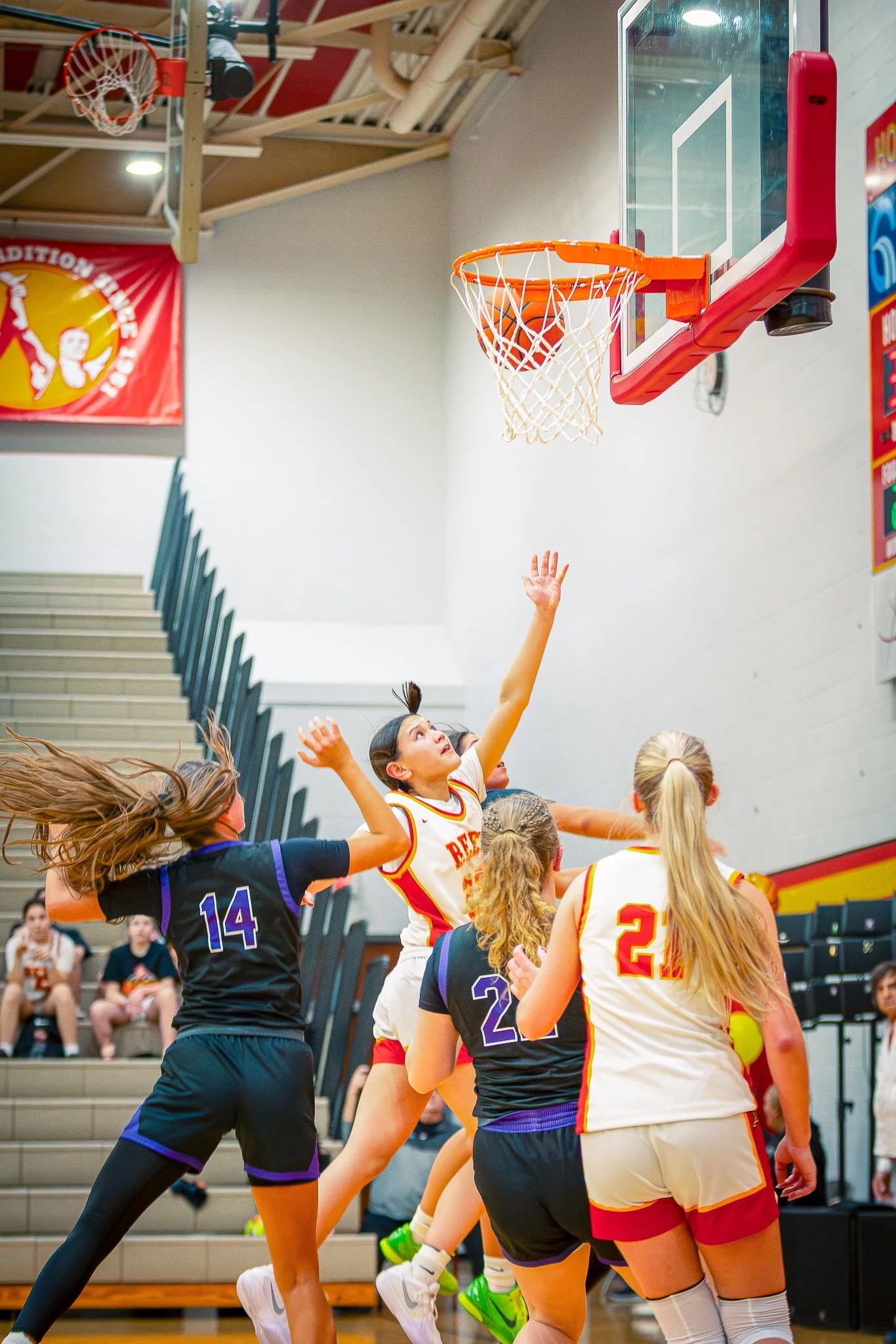 A girls' basketball game in action with players jumping and reaching for the ball near the hoop inside a gym with spectators watching.