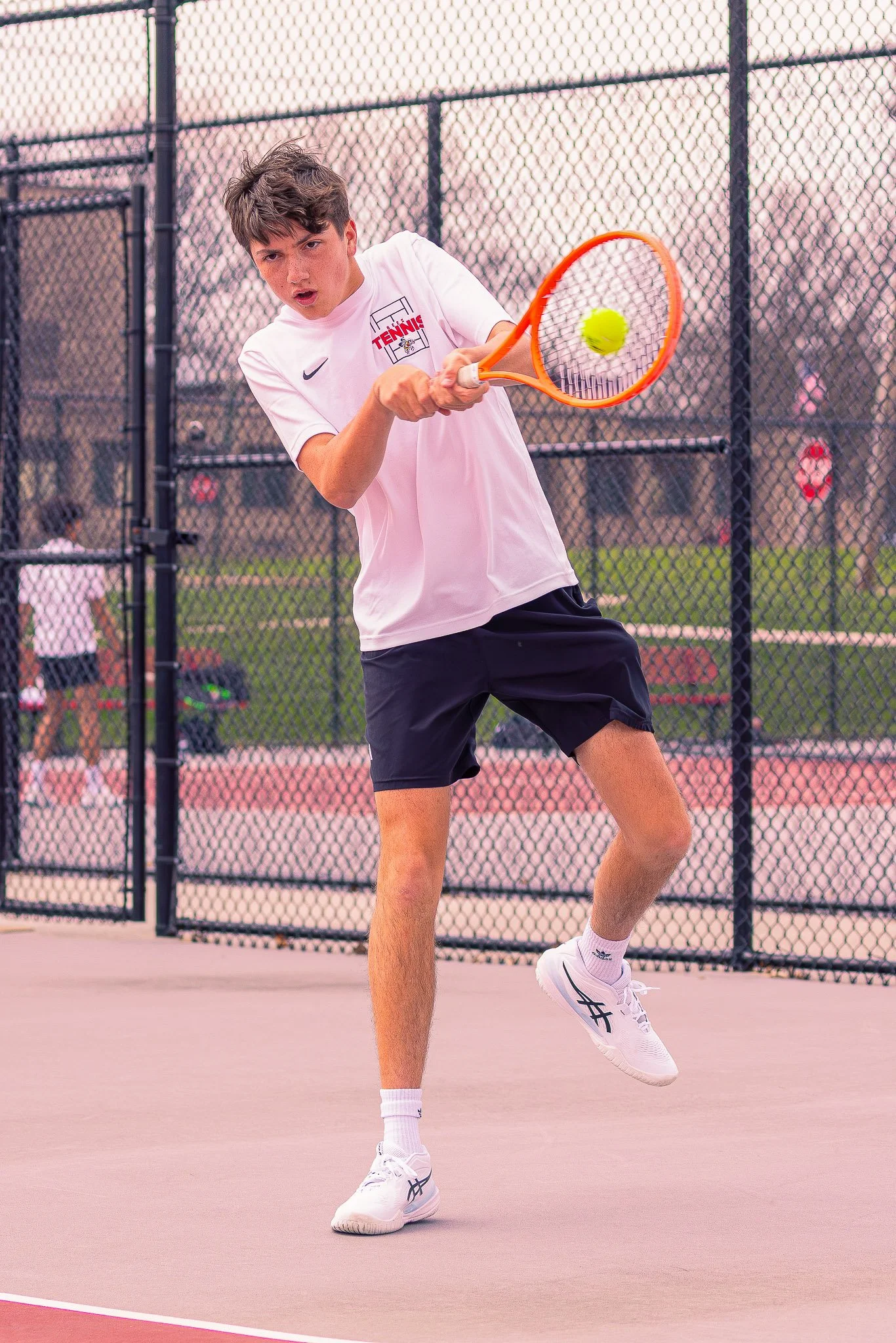 A young man playing tennis on an outdoor court, hitting a yellow tennis ball with an orange racket, wearing a white shirt, black shorts, and white sneakers.