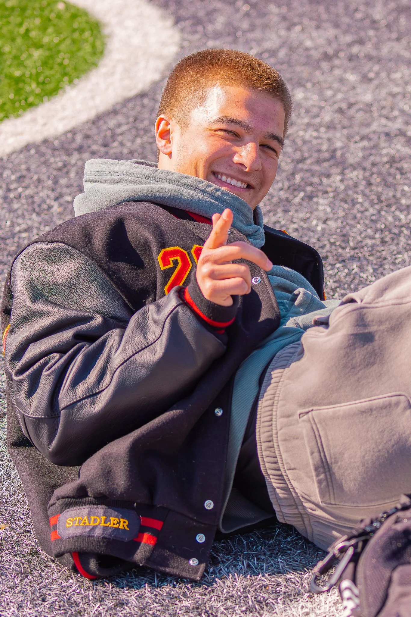 A track runner with short light brown hair smiling and pointing at the camera, wearing a black varsity jacket with red and yellow accents, sitting on purple and white turf outdoors.