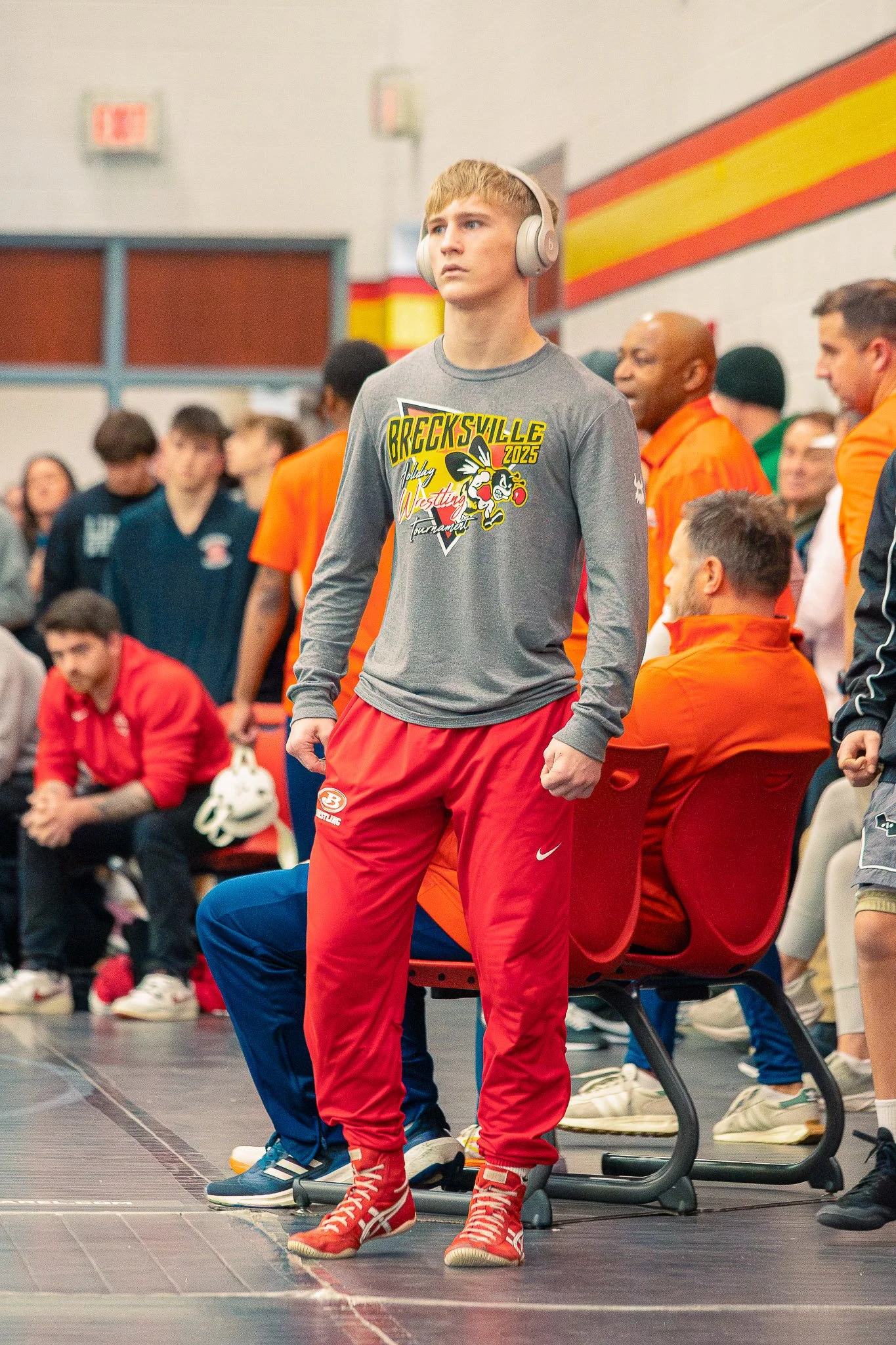 Young man in red pants and orange wrestling shoes standing on an elliptical machine at a wrestling tournament, surrounded by people seated and standing, some in wrestling uniforms and others in casual clothing, in a gymnasium with colorful walls.