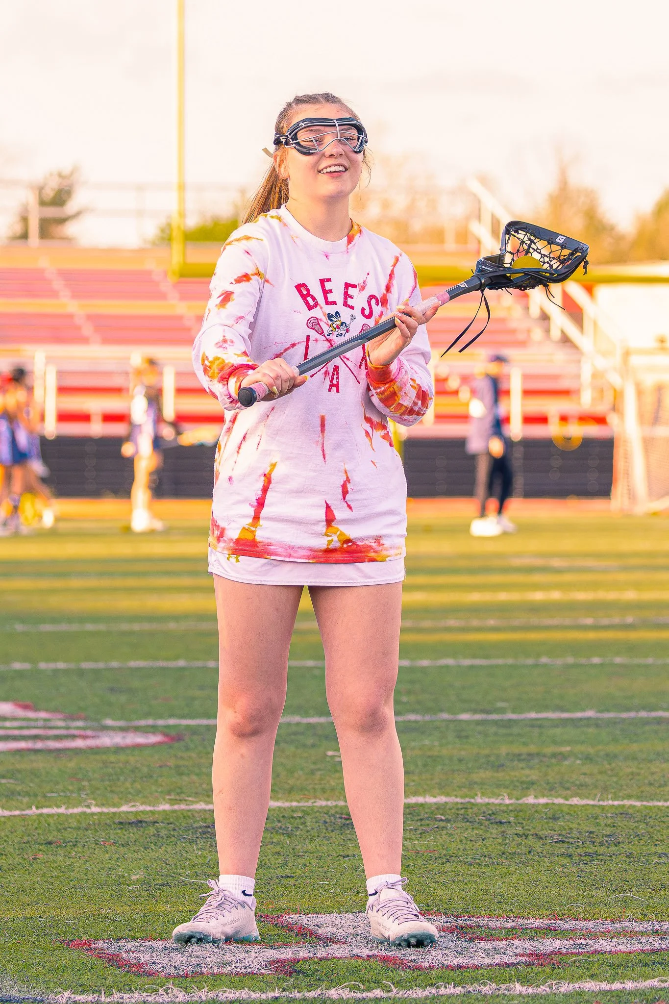 A young woman standing on a sports field holding a lacrosse stick, wearing protective goggles, a white and orange tie-dye long-sleeve shirt, white shorts, and athletic shoes. The background shows empty bleachers and other people on the field.