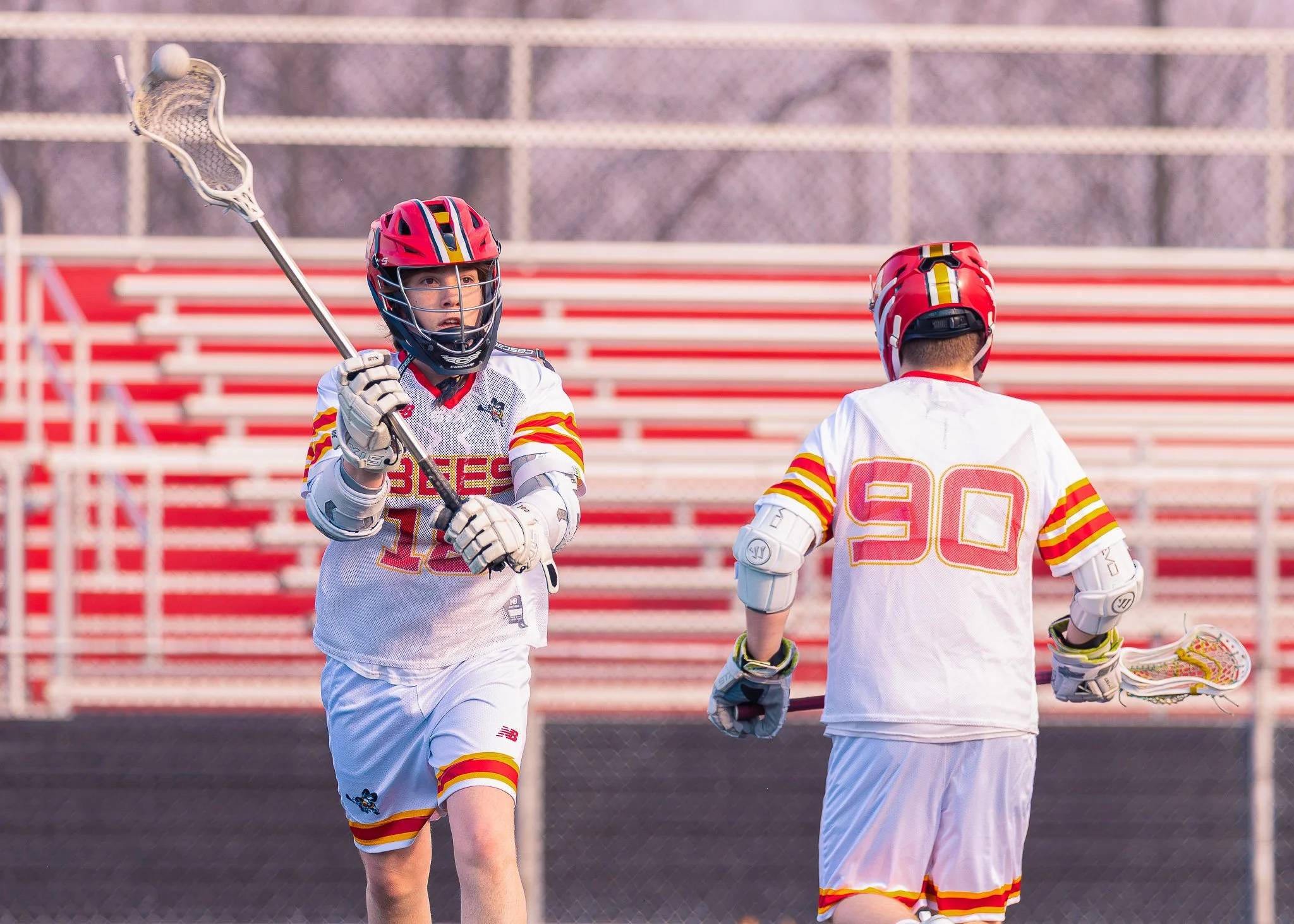 Two young lacrosse players in white jerseys with red and yellow trim on a lacrosse field, wearing helmets and gloves, one holding a lacrosse stick.