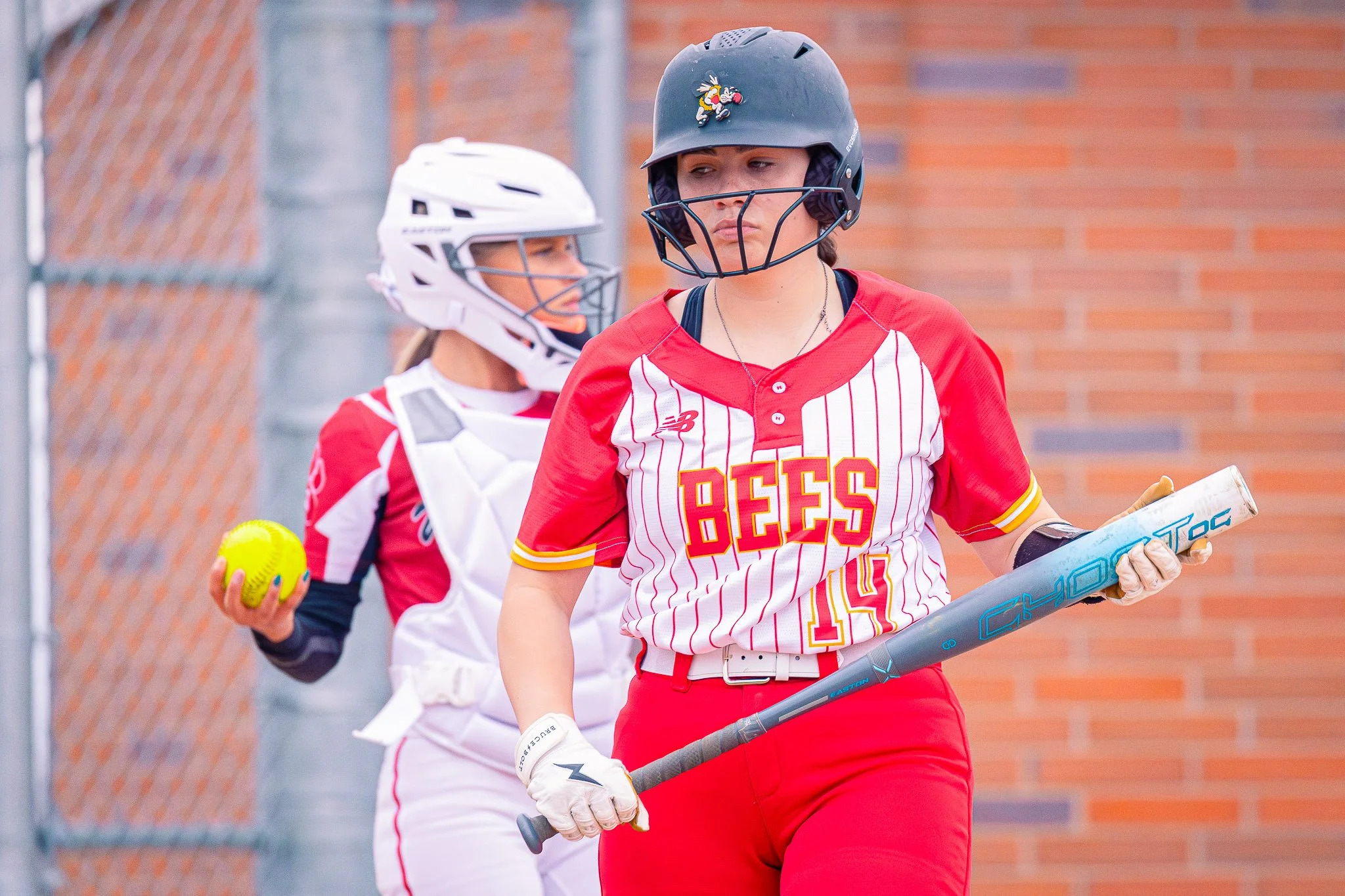 Two female softball players wearing helmets and gloves stand near a chain-link fence, one holding a softball and the other holding a bat. The player in front is wearing a red and white uniform with the word "BEES" on her jersey.