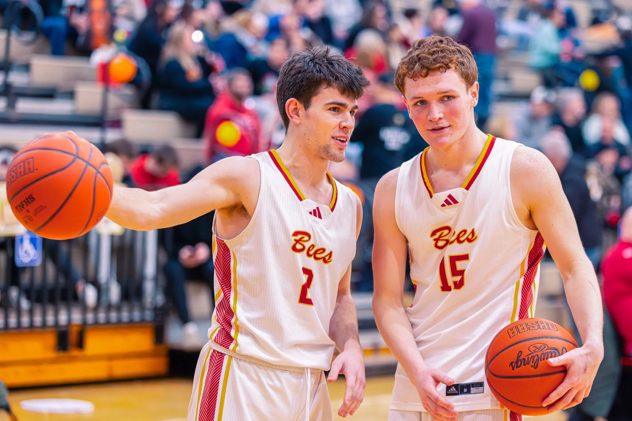 Two basketball players in white jerseys with red and yellow accents, standing on a court, with one holding a basketball, seemingly discussing during a game or practice, surrounded by an audience.