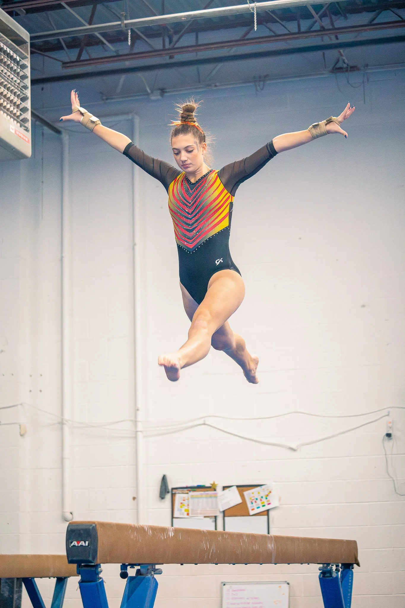 A young female gymnast in a colorful leotard practicing her routine on the balance beam in a gymnasium.