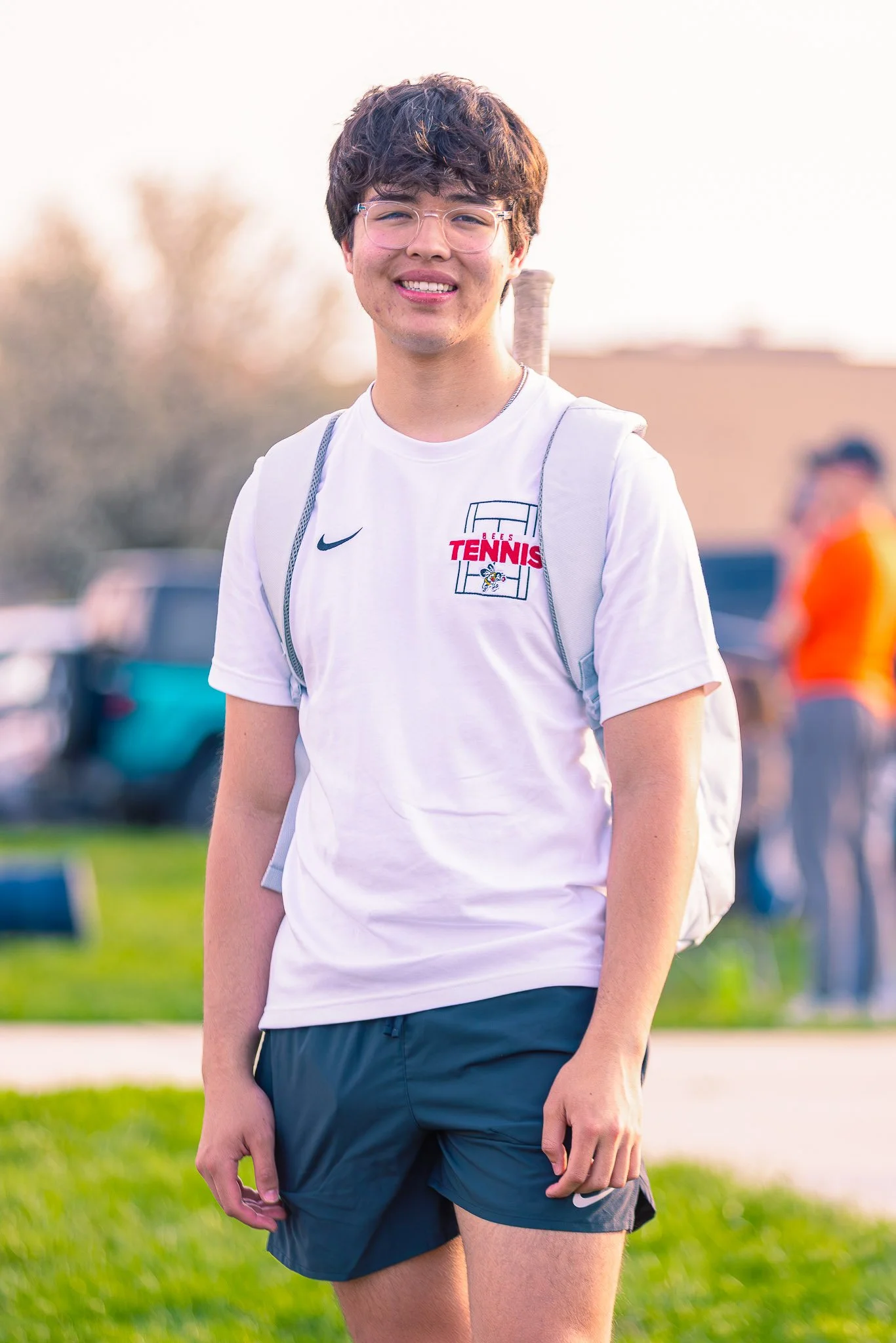 Young man with glasses and curly brown hair smiling outdoors, wearing a white T-shirt with a tennis logo, black shorts, and a backpack, standing on grass with blurred background.