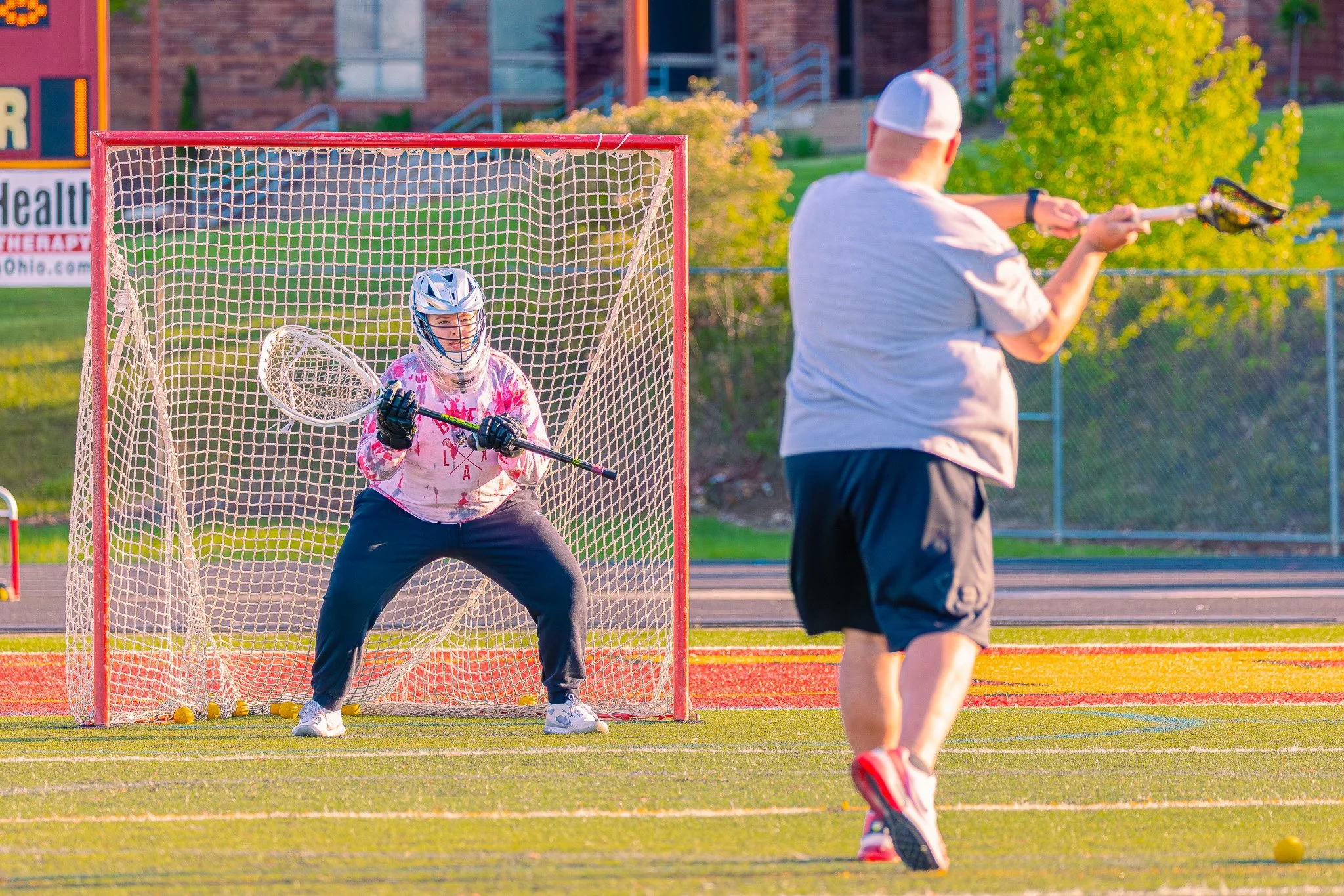 A young girl dressed as a goalie in a pink and white jersey, protective helmet, and gloves, holding a lacrosse stick in front of a lacrosse goal, with a man shooting a ball at her during a game or practice on a grass field in the evening.
