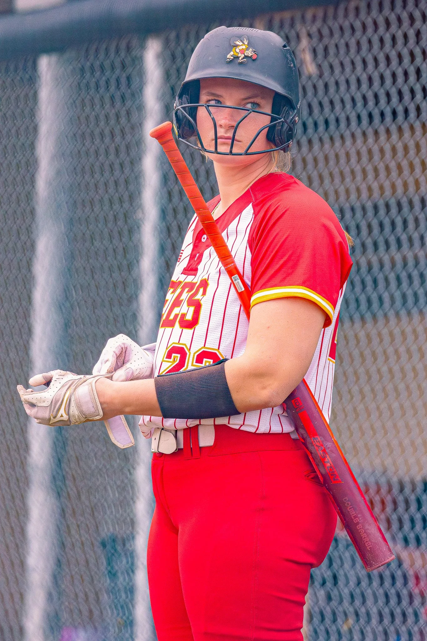 A female softball player in a red and white striped jersey holding a softball bat, wearing a black helmet with a yellow sticker, and standing in front of a chain-link fence.