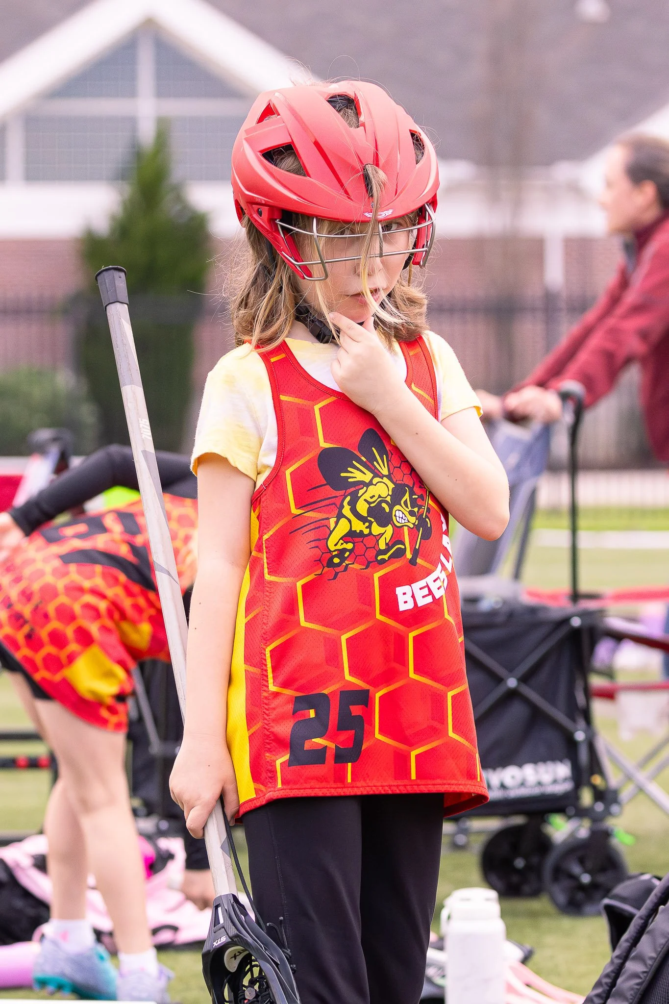 A young girl wearing a red helmet and a red sports jersey with a bee and honeycomb design, standing outdoors, holding a hockey stick. She appears to be preparing for or participating in a sporting event.