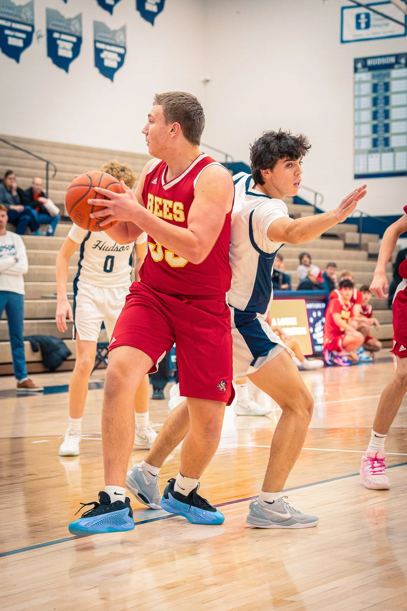 Two young men playing basketball, one in a red jersey and the other in a white and blue jersey, in a gymnasium with spectators on the bleachers.