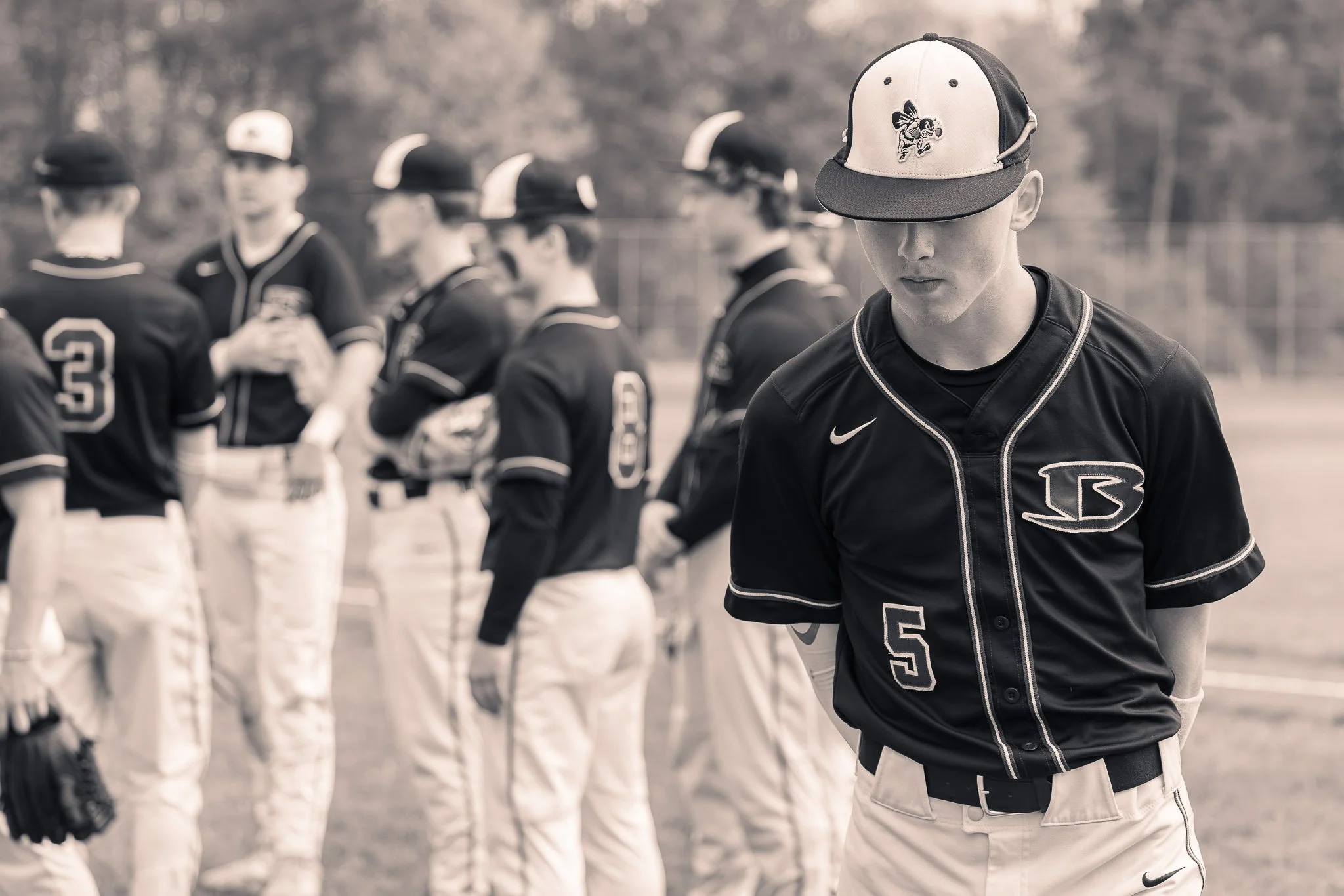 A young baseball player wearing a black jersey with the number 5 and a baseball cap, looking down with a serious expression. In the background, other players in similar uniforms are standing in a line on a baseball field.