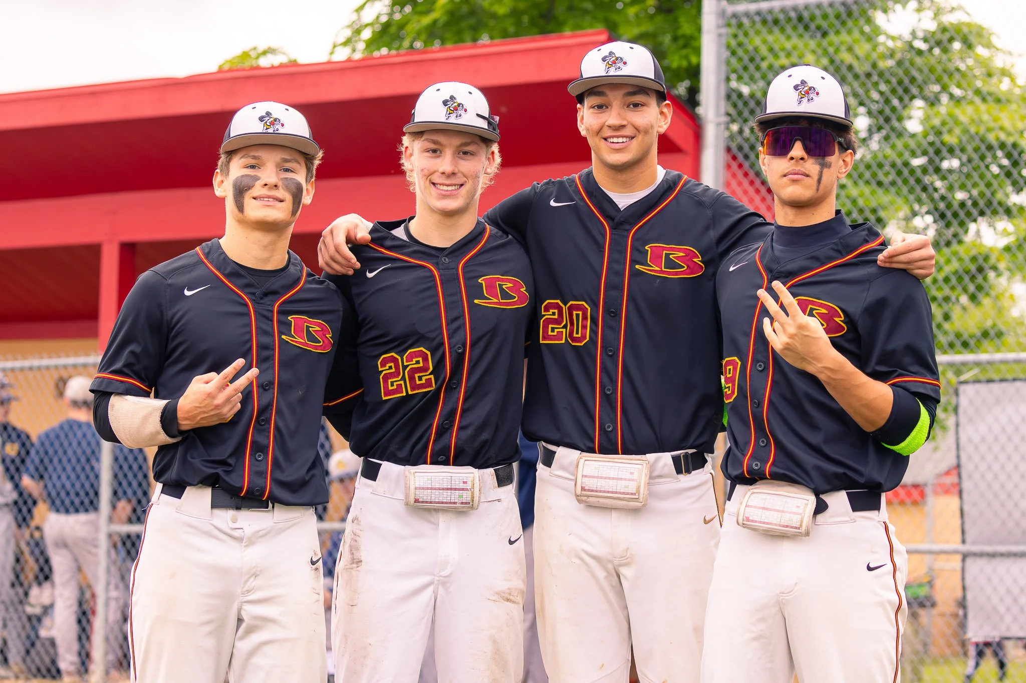 Four young male baseball players in black uniforms with red and yellow accents, standing side by side with arms around each other on a baseball field. They are wearing gray caps with a logo of a bee, and two players are wearing face paint. The backgr