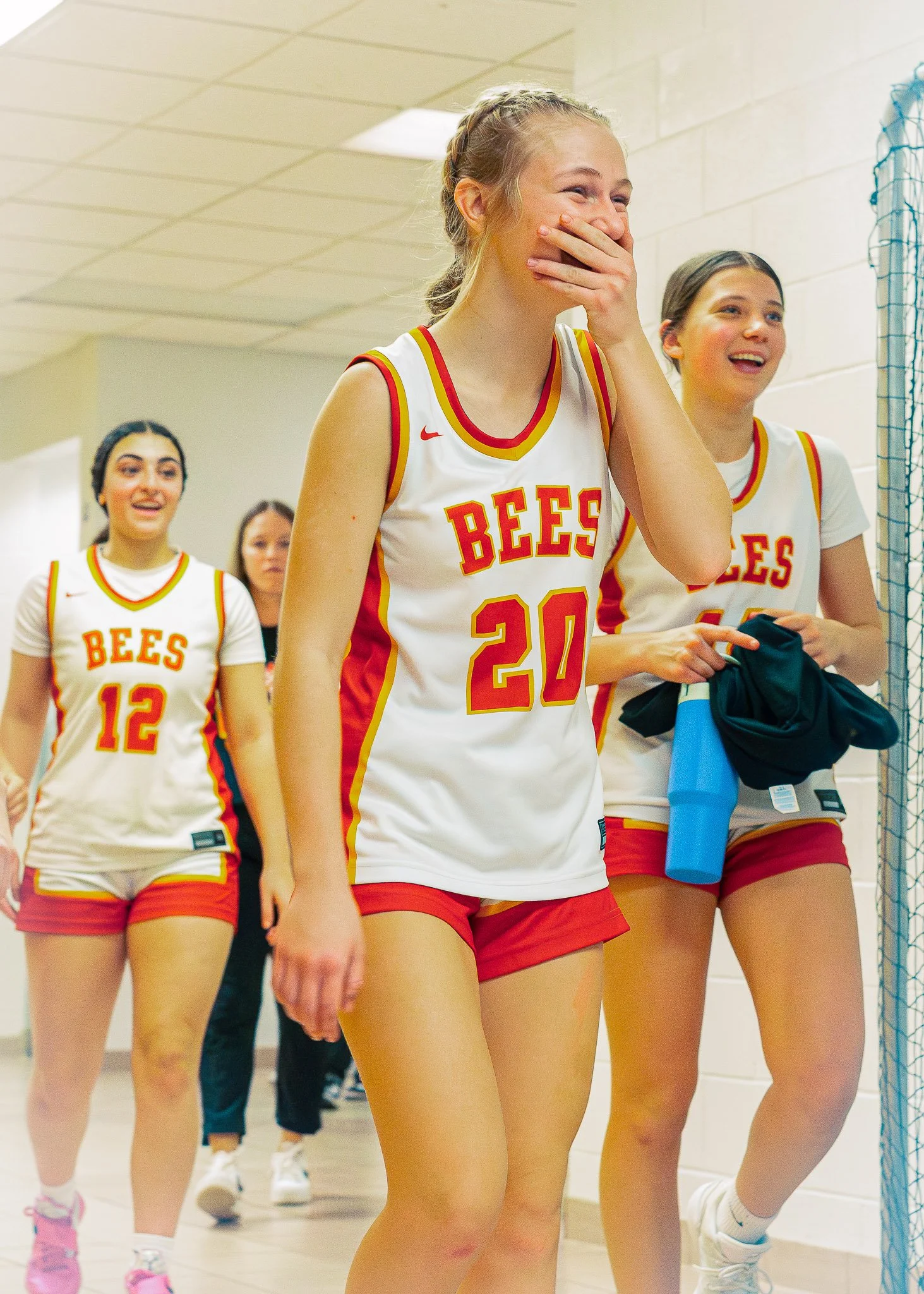 A group of young female basketball players in white jerseys with red and yellow accents, walking indoors, some smiling and laughing.