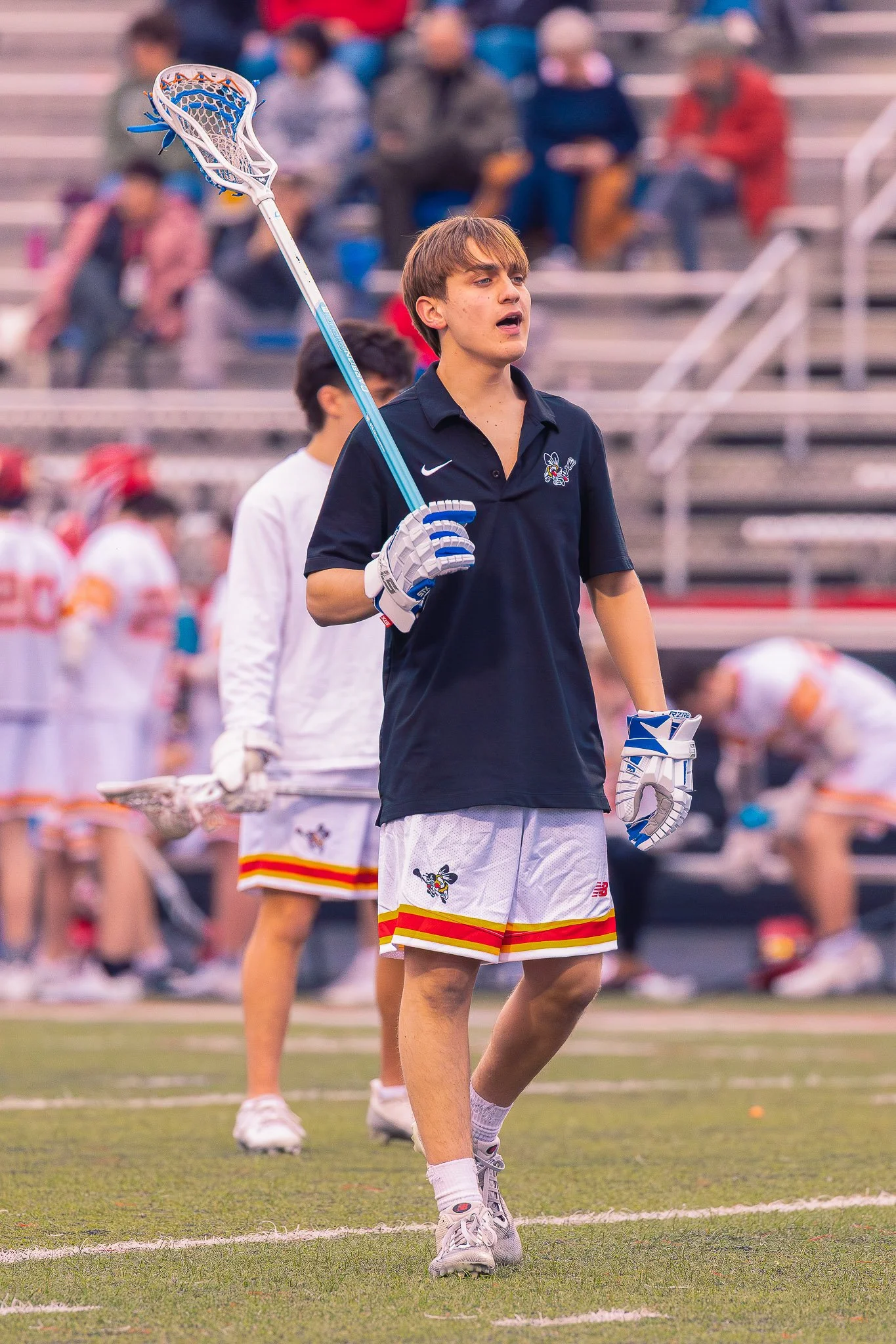 A young male lacrosse player in a navy blue jersey and white shorts holding a lacrosse stick on a field during a game. There are spectators seated in the background.