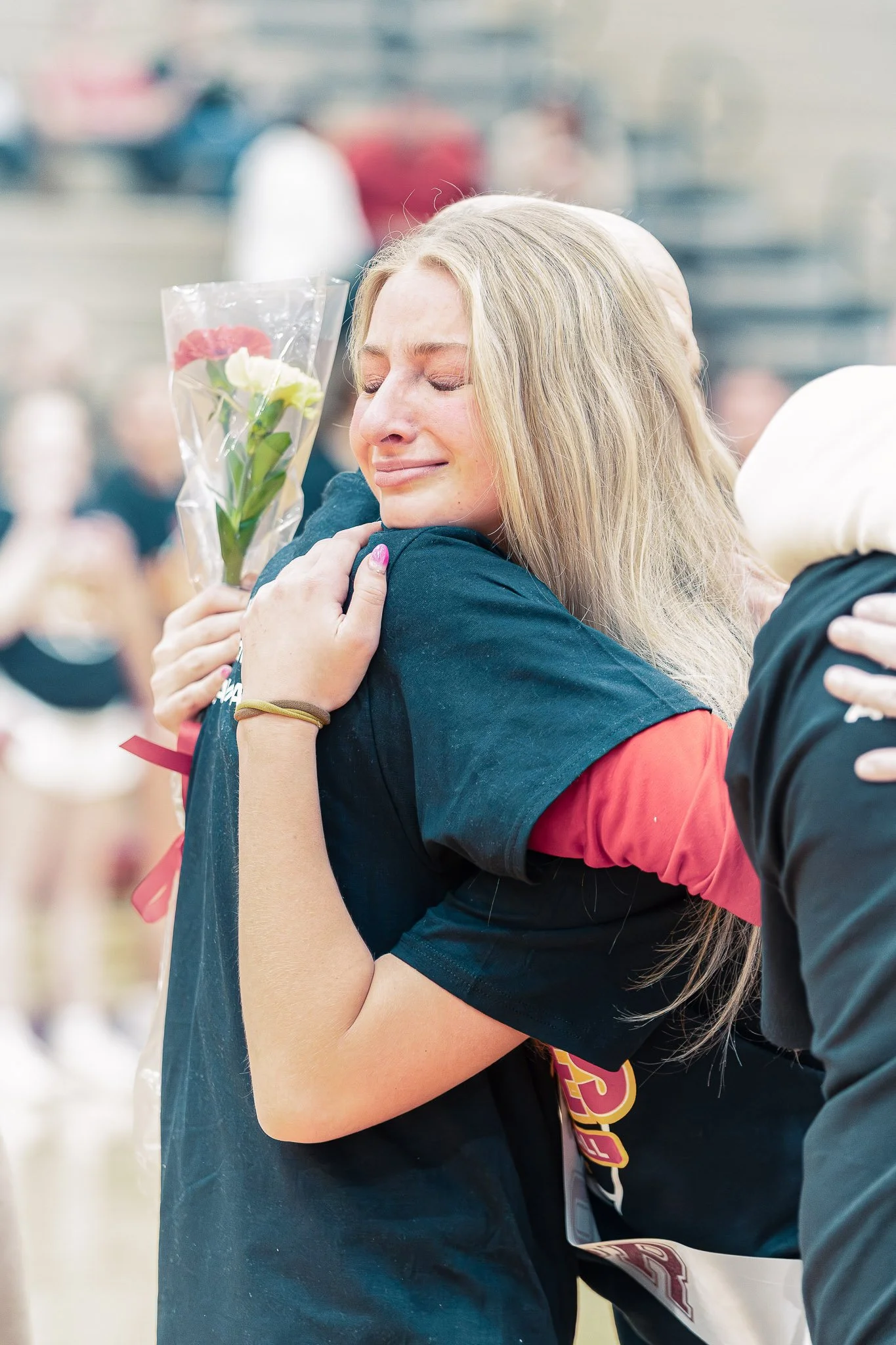 A woman with blonde hair is being embraced by someone with a dark shirt and holding a bouquet of flowers. The woman has a tearful smile and appears to be emotional, possibly crying. The background is blurred but suggests a public or outdoor setting w