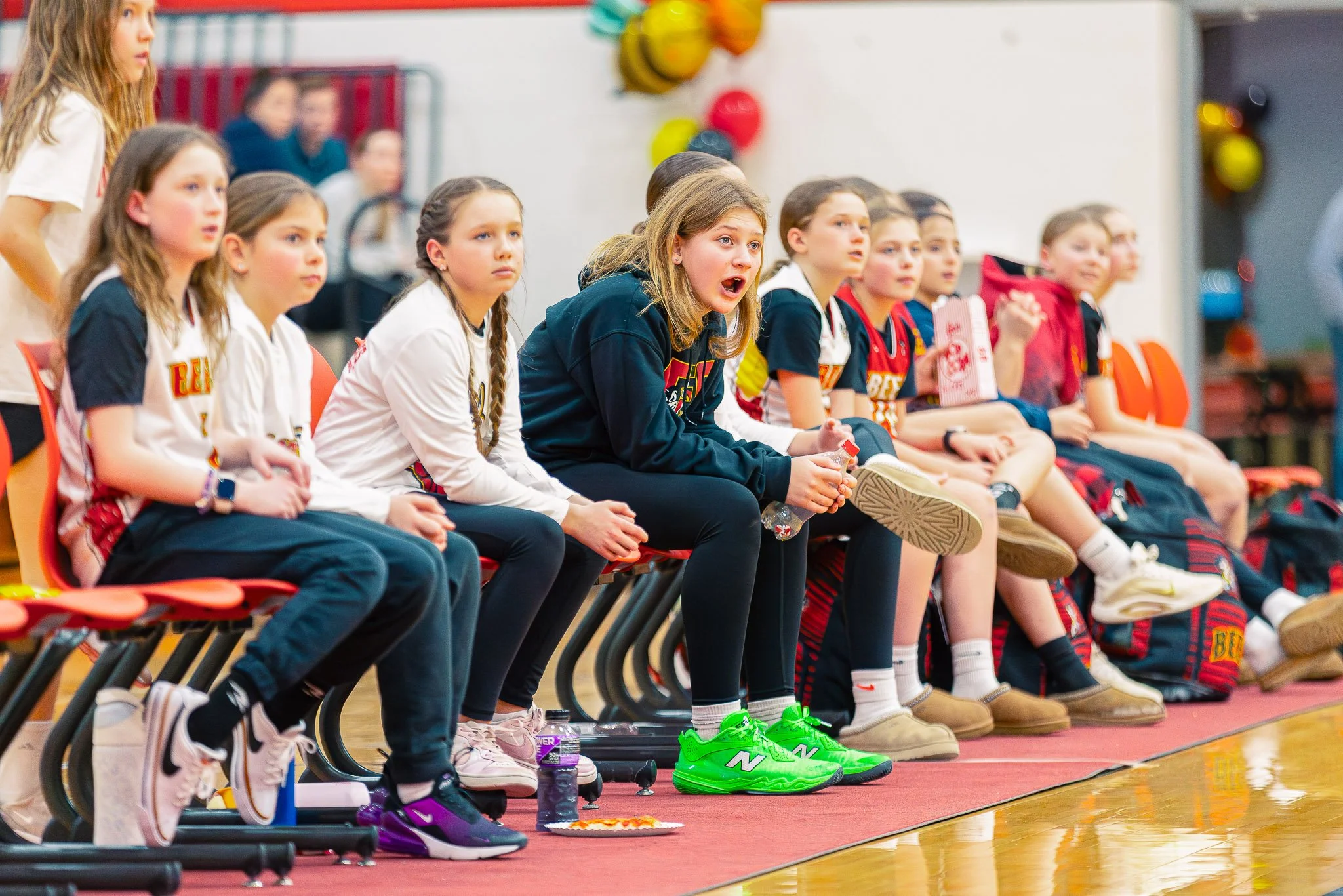 Group of young girls sitting on chairs, watching an event attentively in a gymnasium. Some girls are wearing sports jerseys, sneakers, and casual clothes.